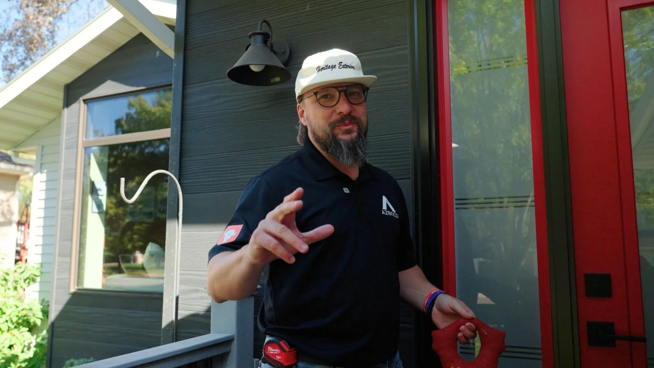 Man standing outside a house with black and red siding, wearing glasses, a white cap that says 'Heritage Exterior,' and a black polo shirt with a logo. He has a beard and is holding a red cloth or towel in his left hand while gesturing with his right hand.