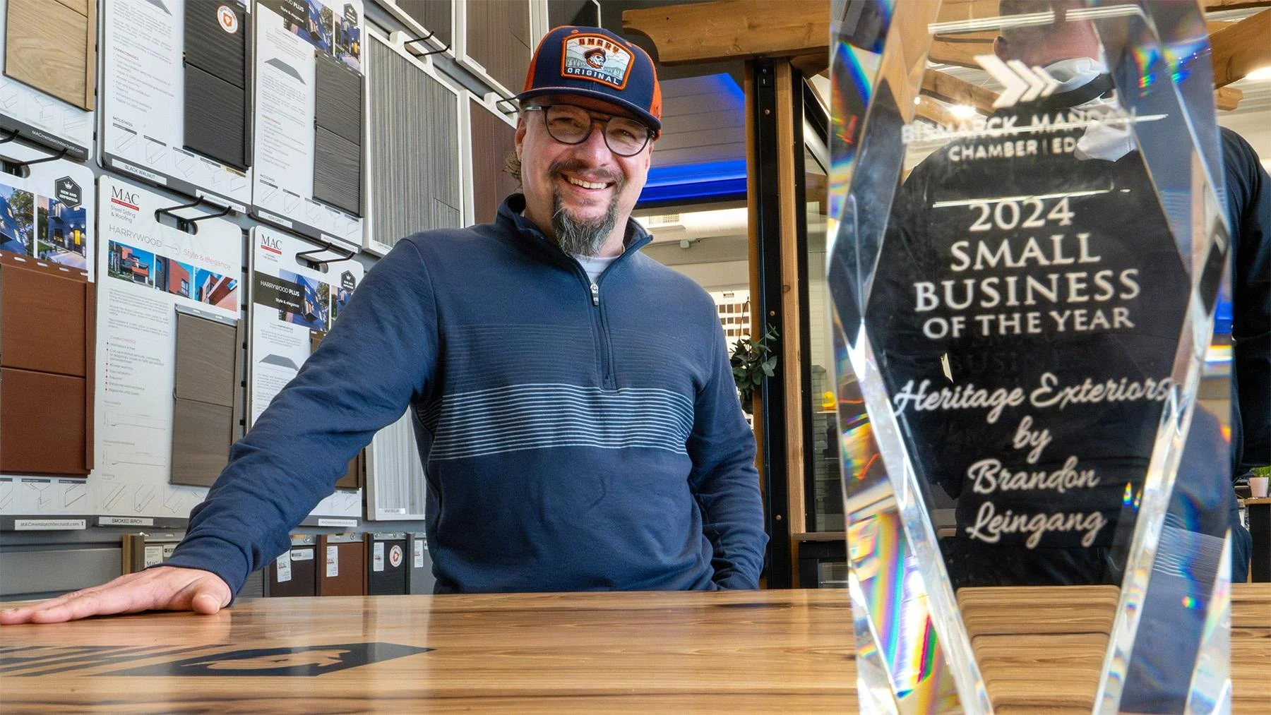 A man with glasses and a beard, wearing a dark blue jacket and a black, orange, and blue baseball cap, smiling while standing behind a wooden table with an award on it. The award reads "2024 Small Business of the Year" and is from Heritage Exterior by Brandon Leingang. The background has displayed samples of siding material on a wall.