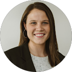 A young woman with long brown hair smiling, wearing a black blazer and a gray top, against a light background.