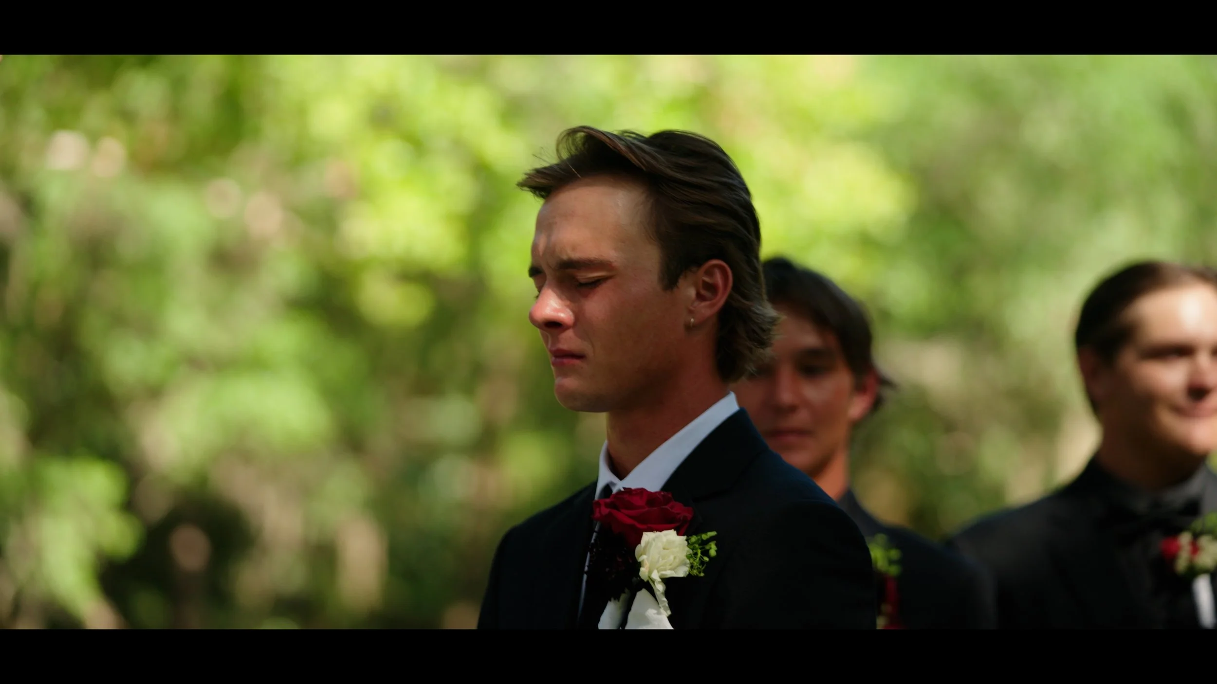 A young man in a tuxedo standing outdoors, with two other men in tuxedos behind him, during a wedding ceremony in a wooded area.