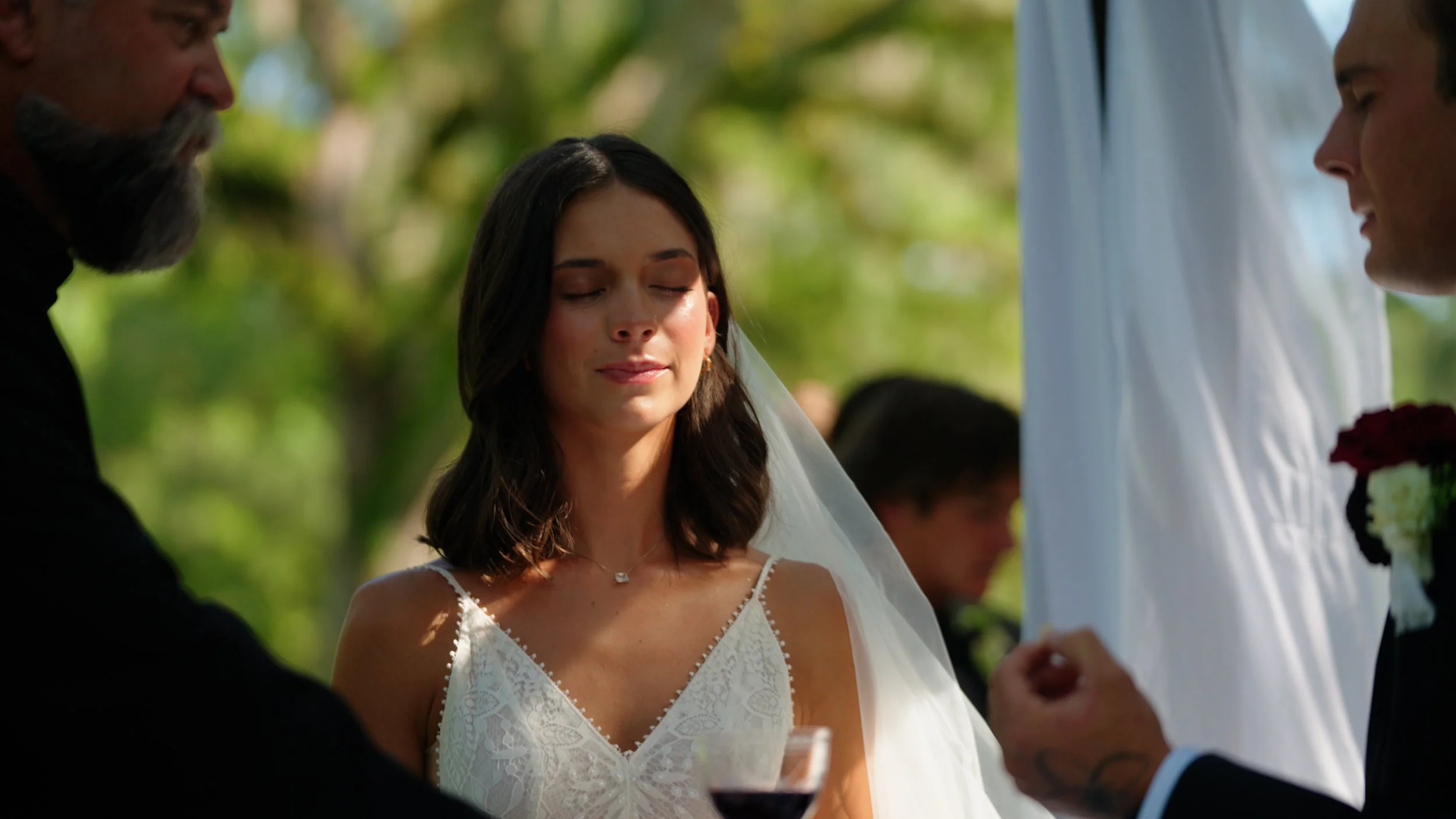 A bride with brown hair, wearing a white lace wedding dress and veil, has her eyes closed during her wedding ceremony outdoors, surrounded by officiant and groom.