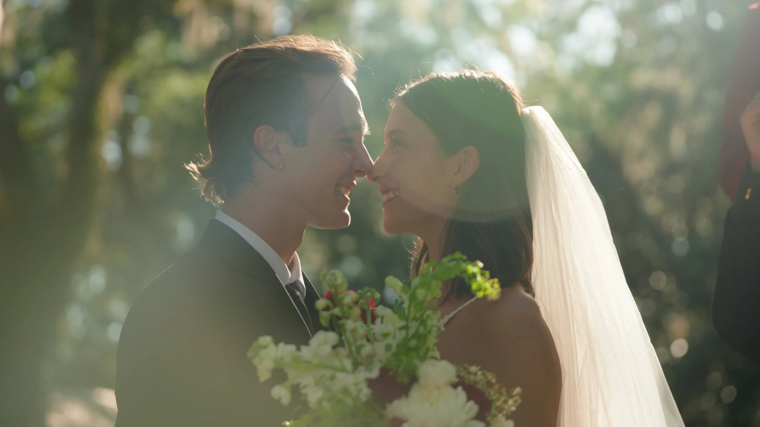 A bride and groom smile and touch noses during their outdoor wedding, with sunlight filtering through trees in the background.
