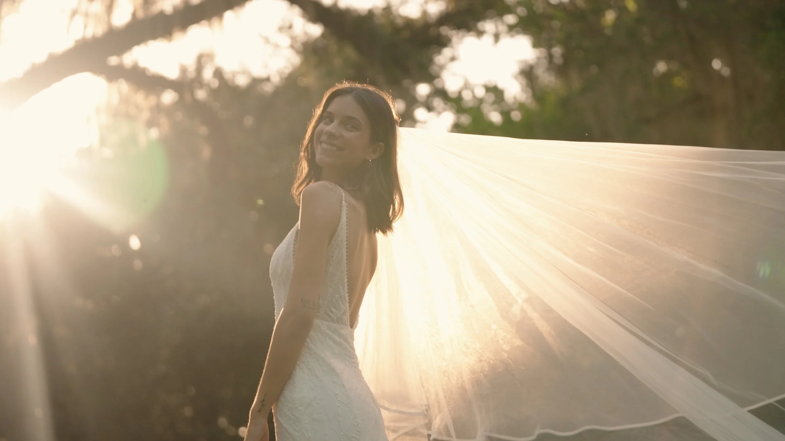 A woman in a white wedding dress with a veil standing outdoors at sunset, smiling and looking back over her shoulder.