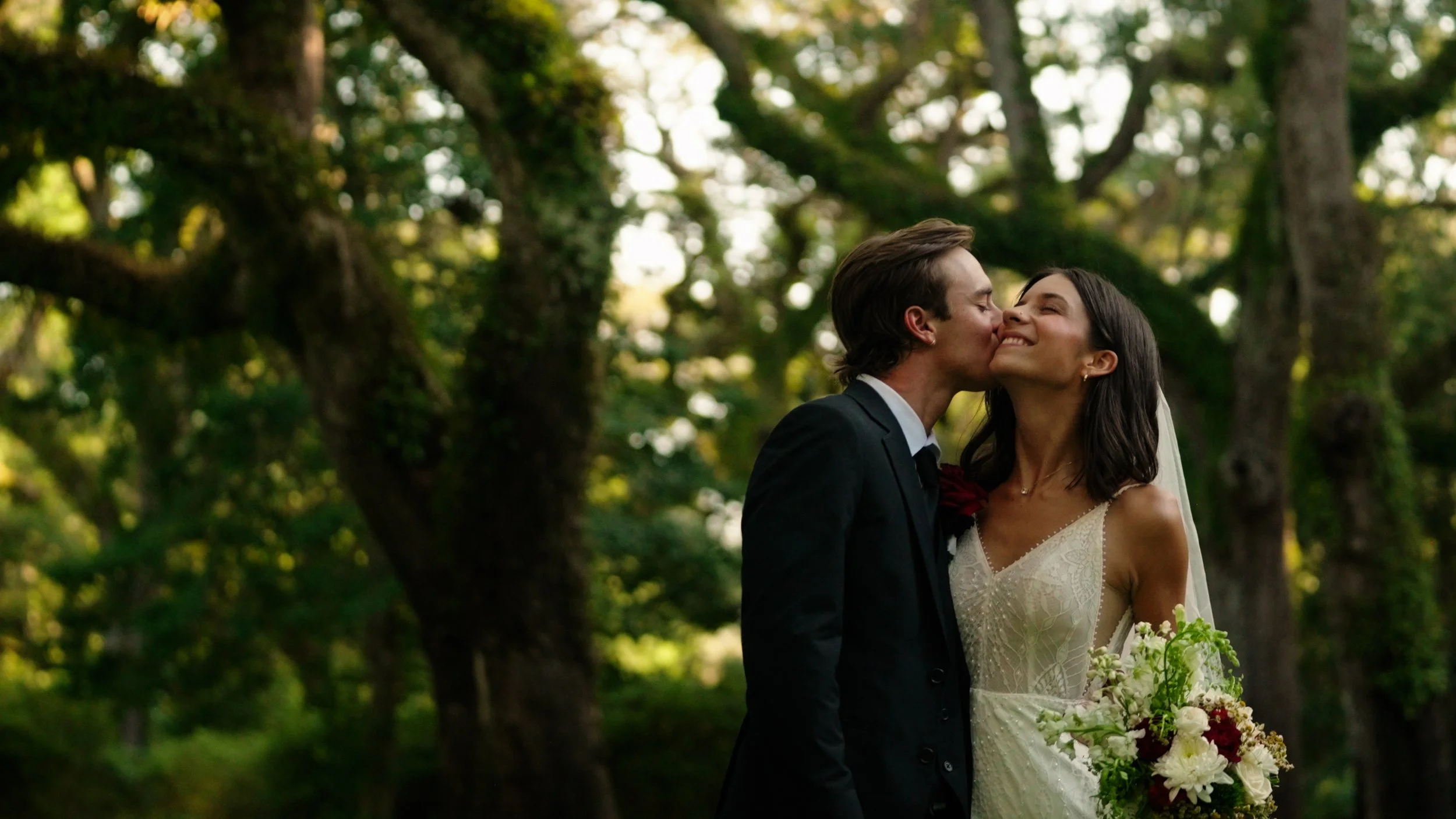 A newlywed couple sharing a kiss outdoors with trees in the background. The bride is in a lace wedding dress holding a bouquet, and the groom is in a black suit with a white shirt and black tie.