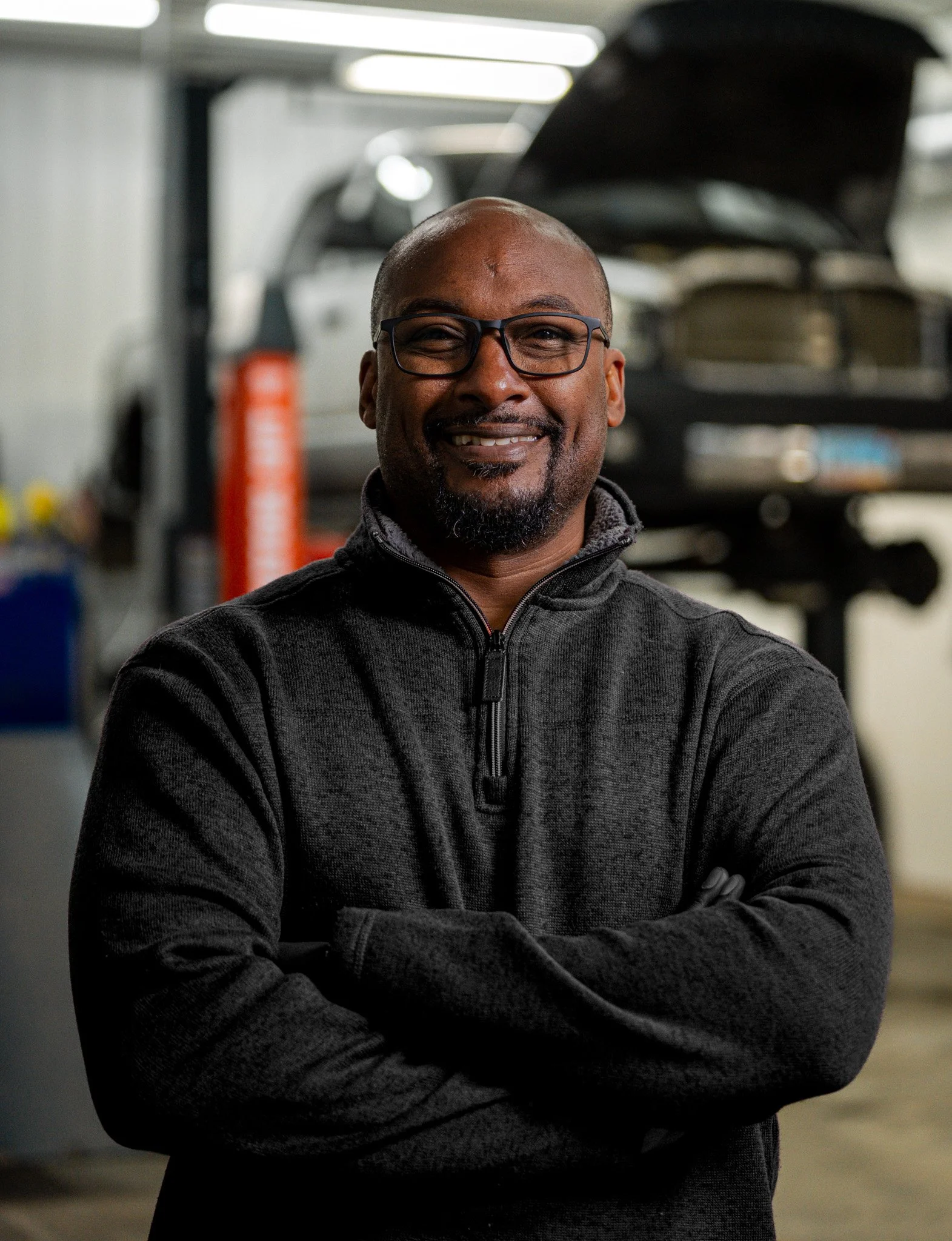 A man with glasses smiling and crossing his arms inside an auto repair shop, with a car on a lift in the background.