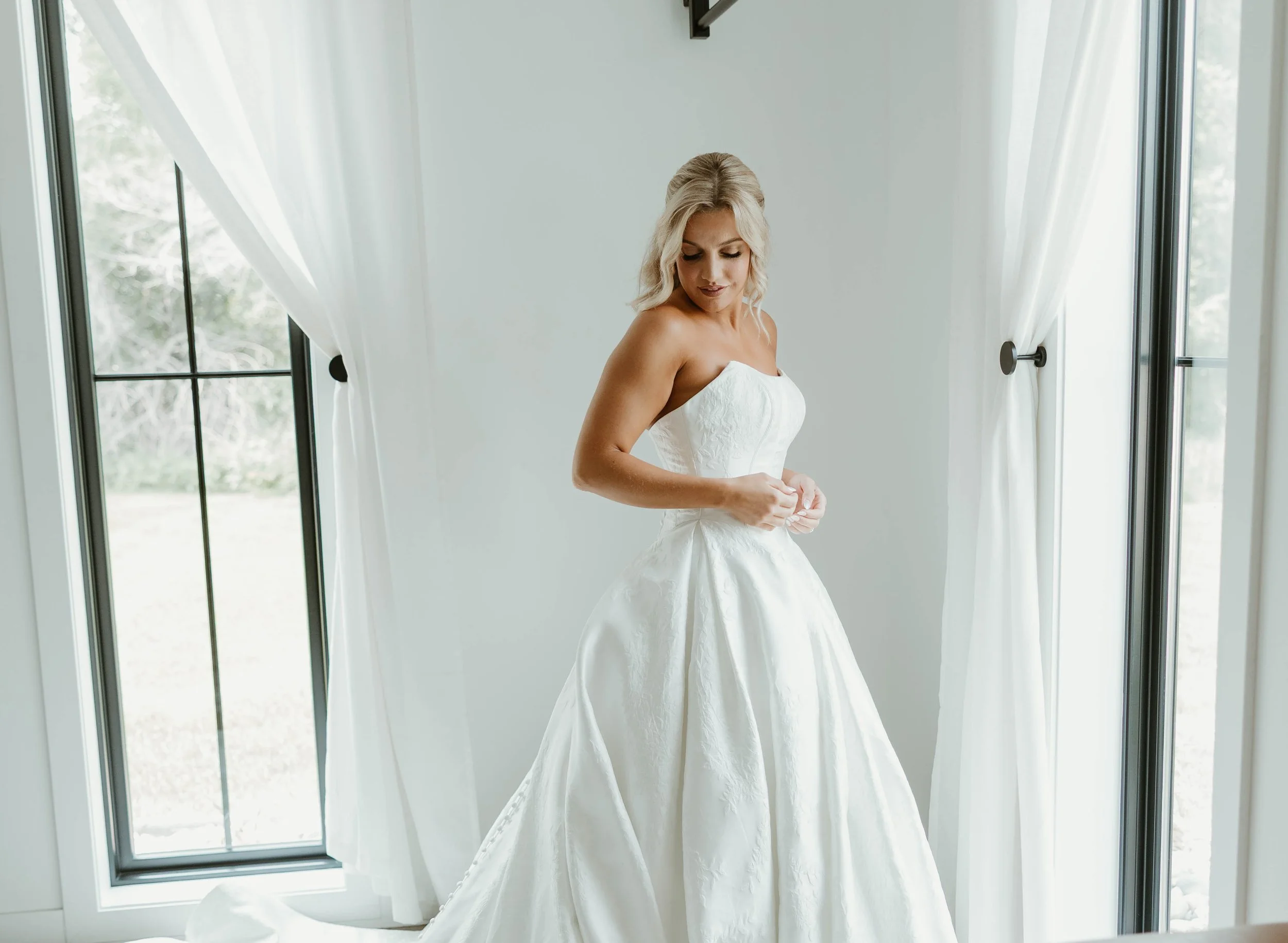 A woman in a strapless white wedding gown standing in front of a large window with white curtains, looking down and adjusting her dress.