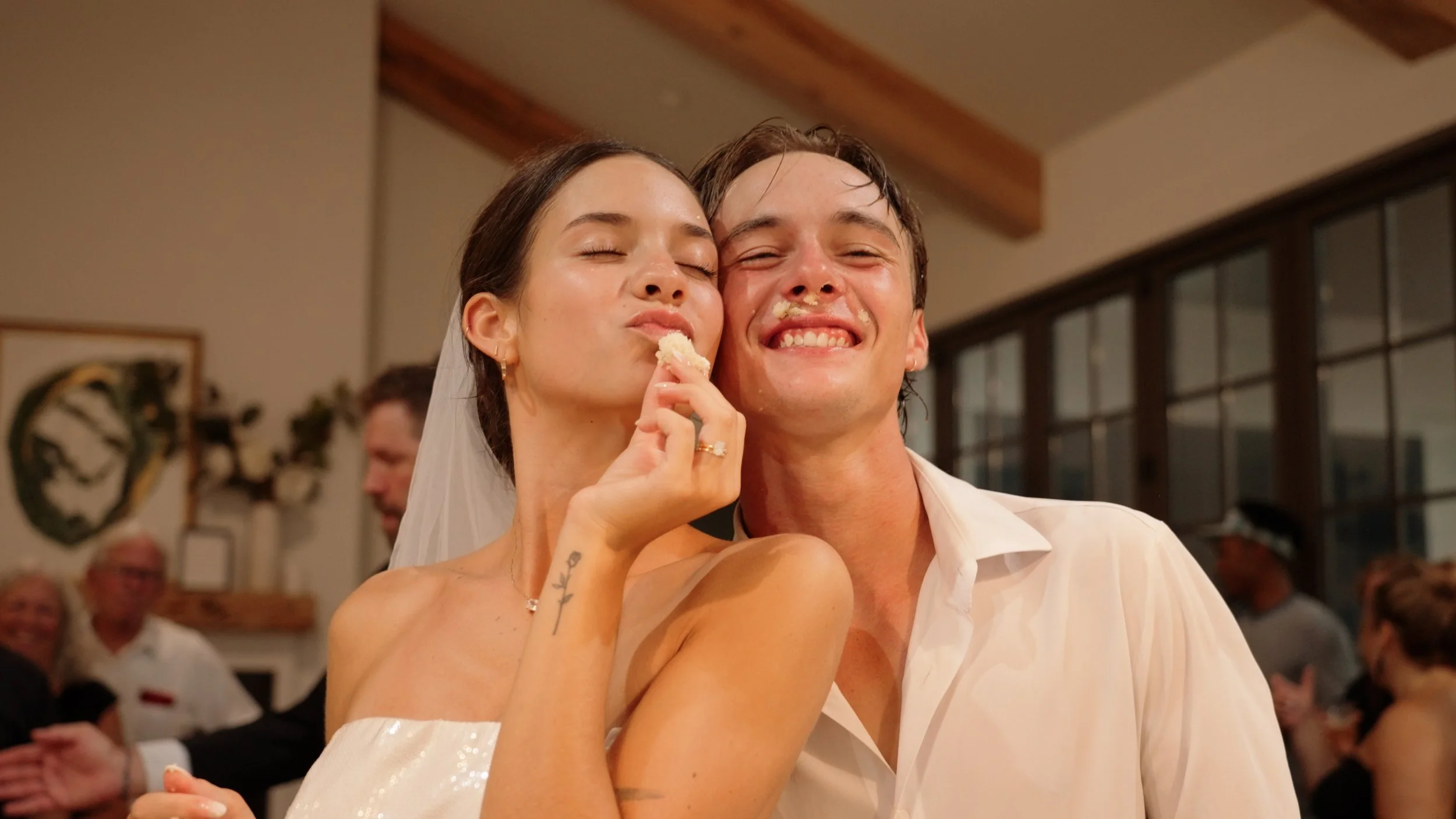 A bride and groom smiling and celebrating at their wedding reception, with the bride holding a piece of cake near her face and the groom with cake on his face.