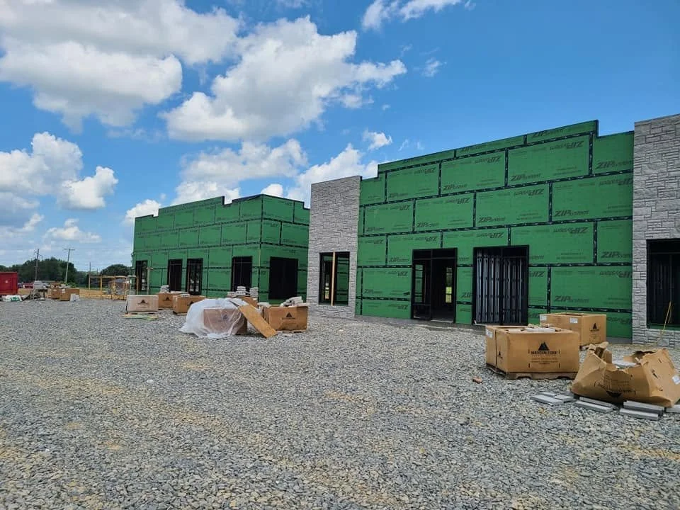 Commercial building under construction with green sheathing, stone veneer, construction materials, and gravel surrounding it