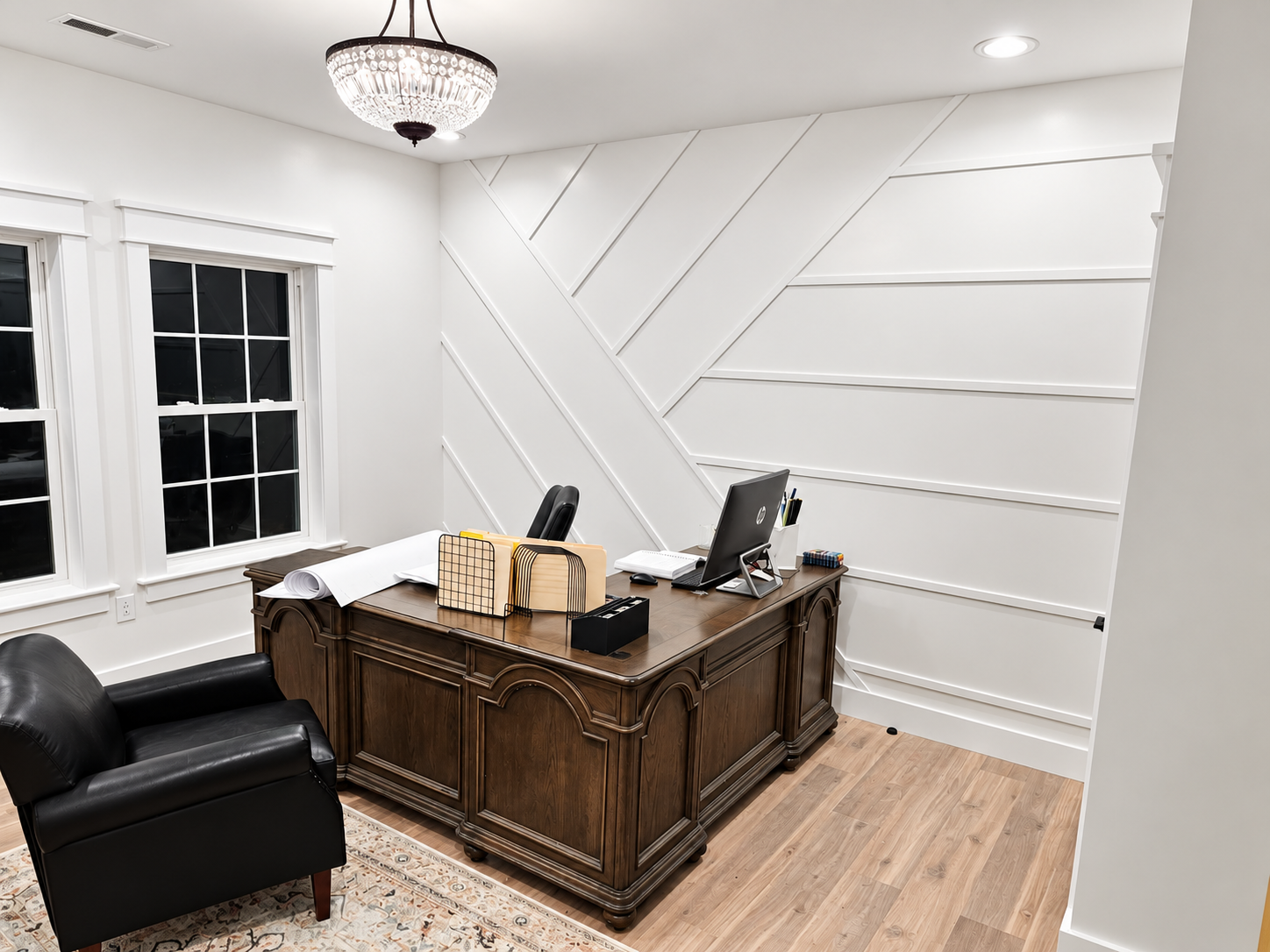 Elegant home office with an ornate wooden desk, laptop, black leather chair, geometric white accent wall, and chandelier