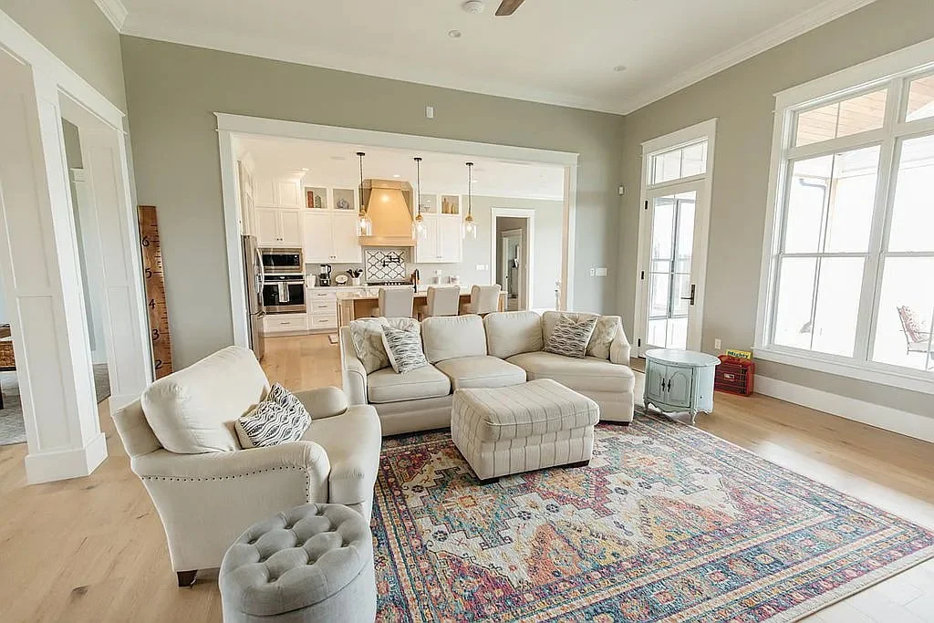 Bright open-concept living room with cream sofas, patterned area rug, ottoman, large windows, and a modern white kitchen in the background.