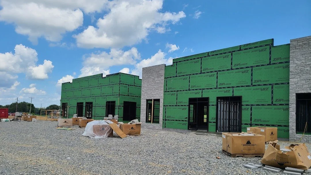 Commercial building under construction with green exterior sheathing, gray stone facade panels, construction materials, and a blue sky with clouds