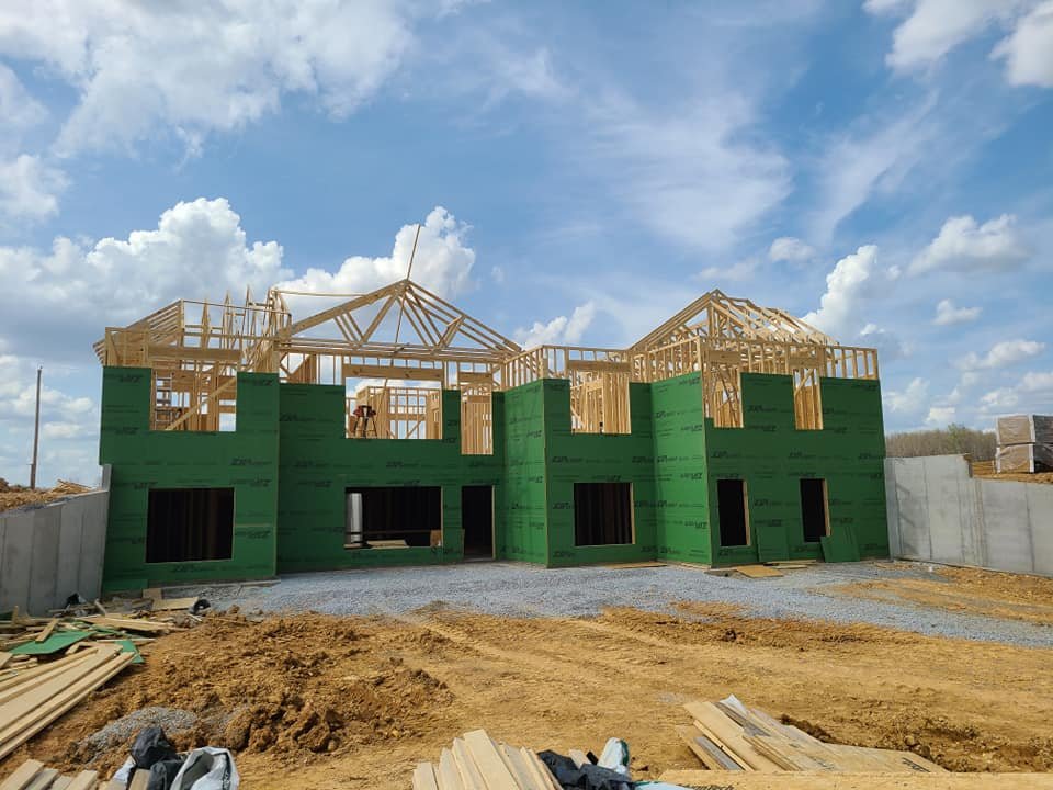 Framed two-story house under construction with green sheathing, exposed roof trusses, and a dirt construction site under a blue sky
