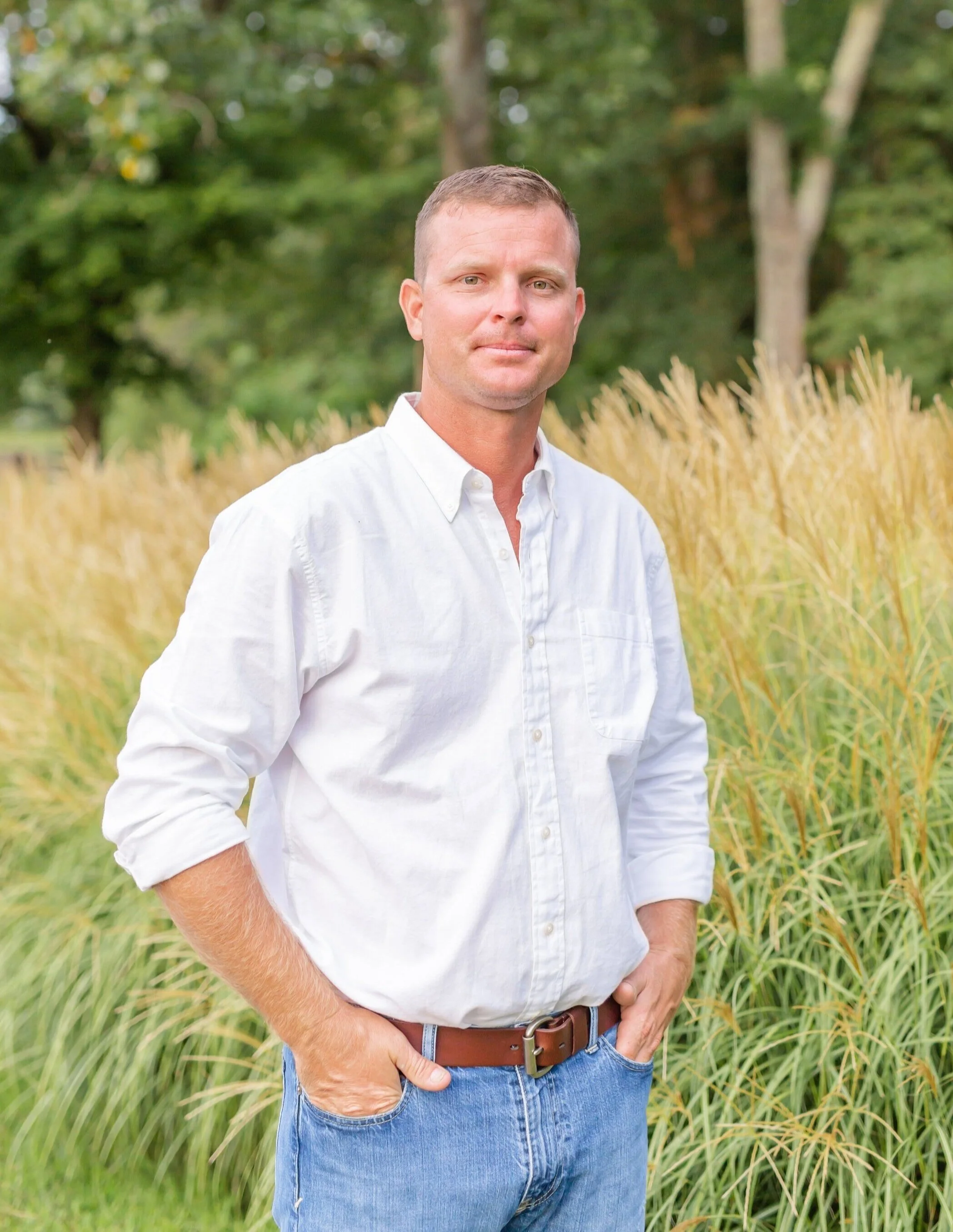 Man in a white button-down shirt and jeans standing in a grassy field outdoors