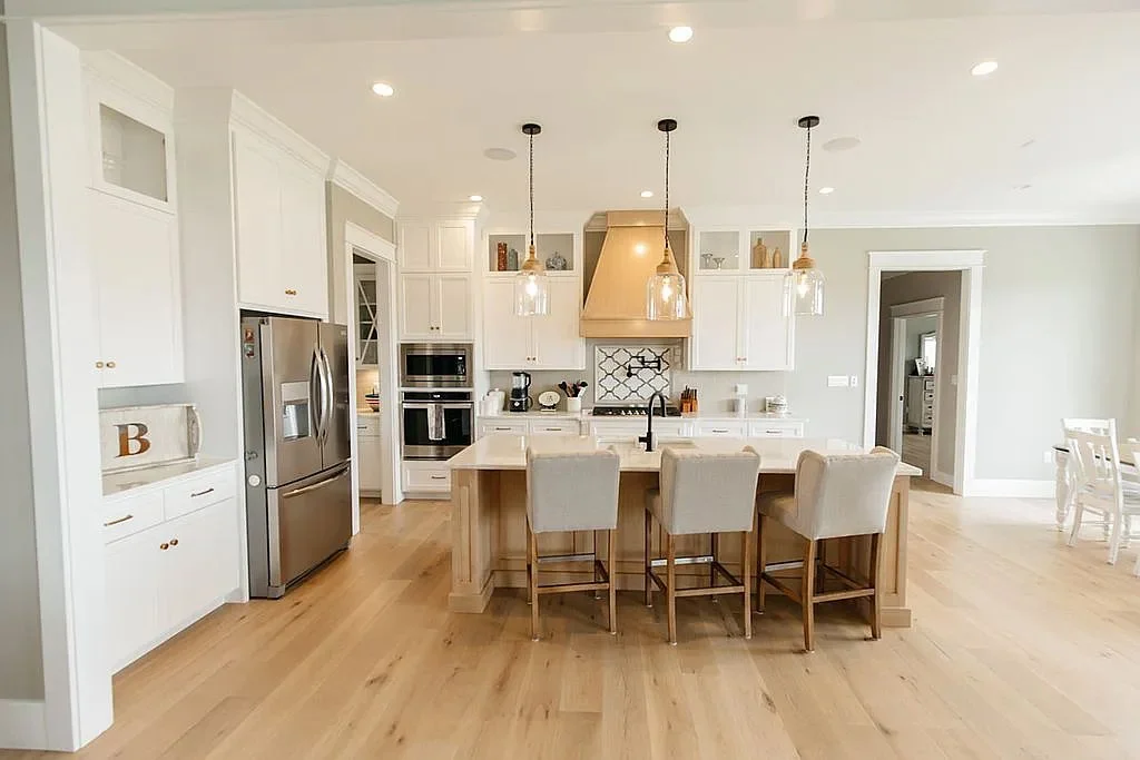 Bright modern white kitchen with a large island, three bar stools, stainless steel appliances, pendant lights, and adjacent dining area