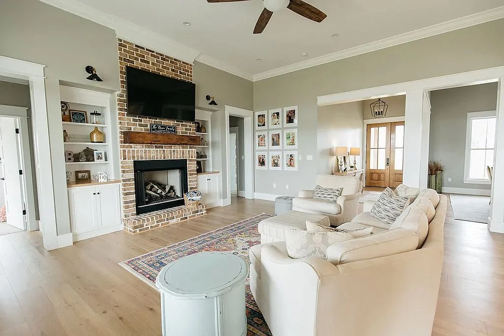 Bright traditional living room with brick fireplace, wall-mounted TV, white sofas, patterned area rug, built-in shelves, and ceiling fan.