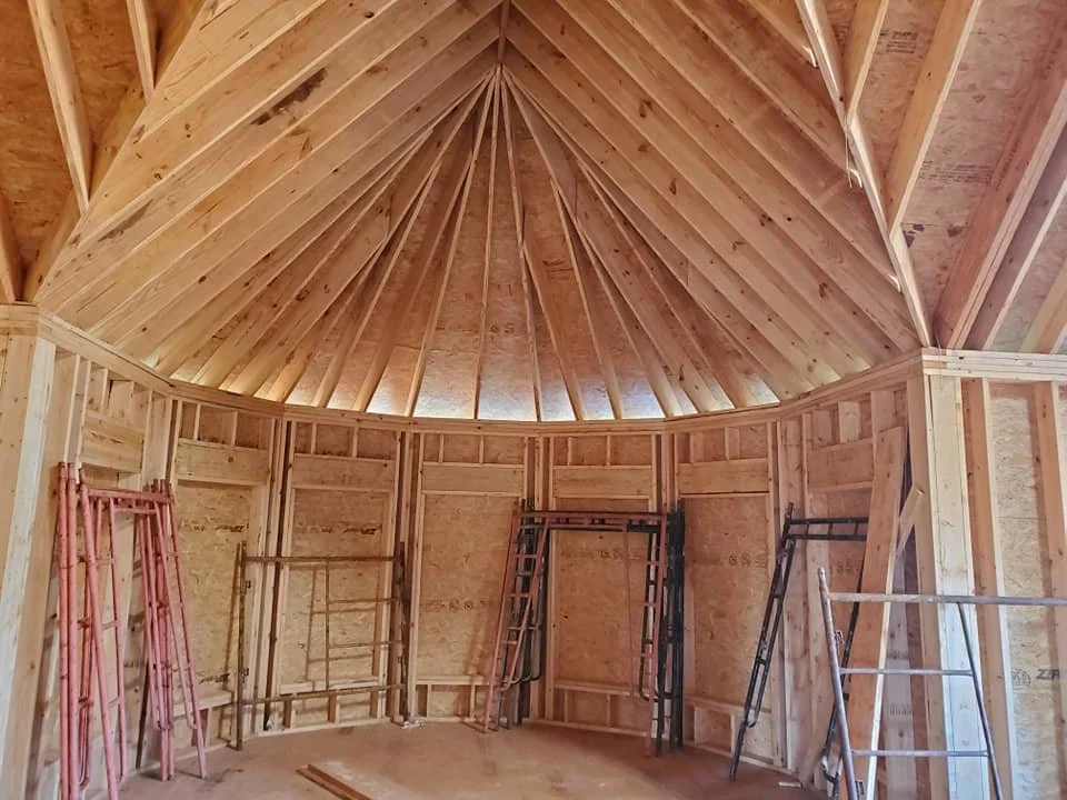 Interior of a partially constructed wooden building with exposed framing, vaulted roof beams, ladders, and construction materials.