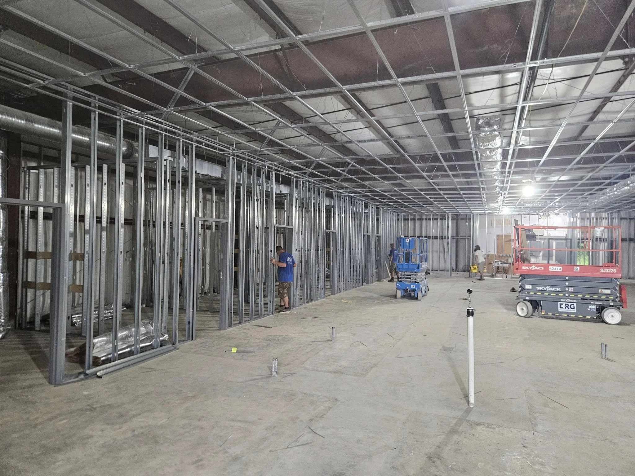 Commercial building interior under construction with metal wall framing, exposed ductwork, workers, and scissor lifts.