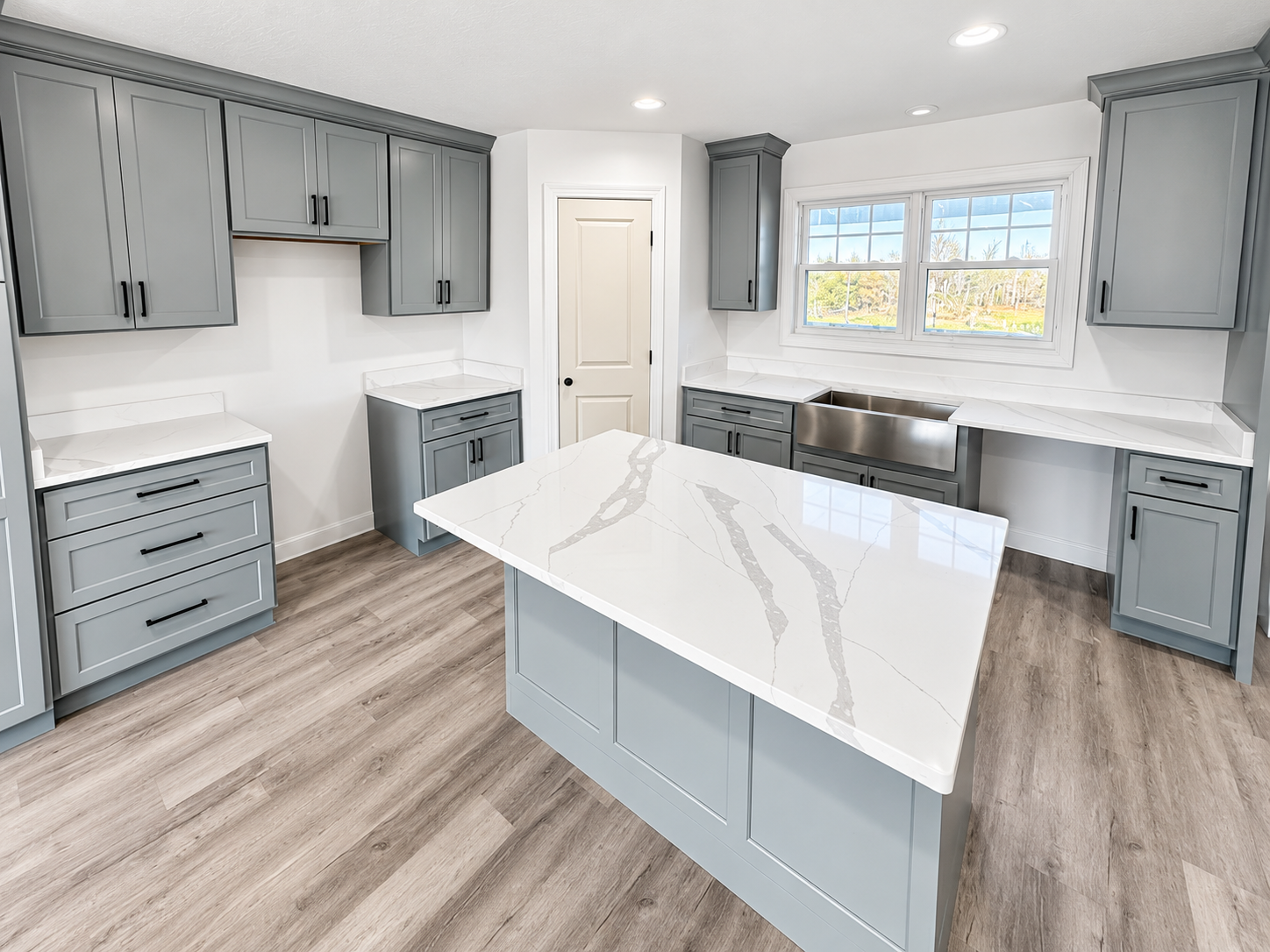 Modern kitchen with gray shaker cabinets, white quartz countertops, large island, farmhouse sink, hardwood floors, and double window