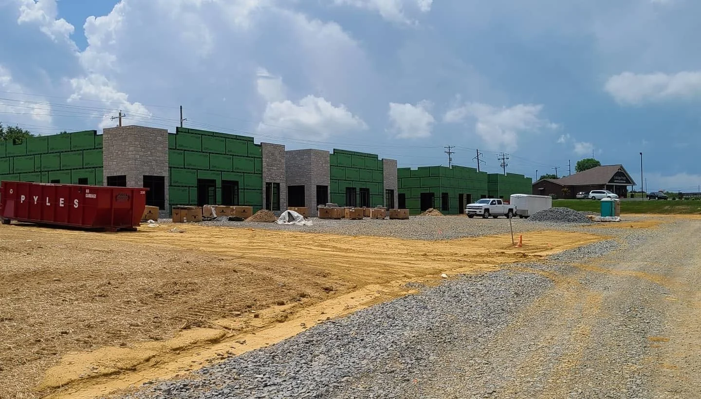 Commercial building under construction with green sheathing, brick columns, construction materials, gravel, and a red dumpster under a partly cloudy sky