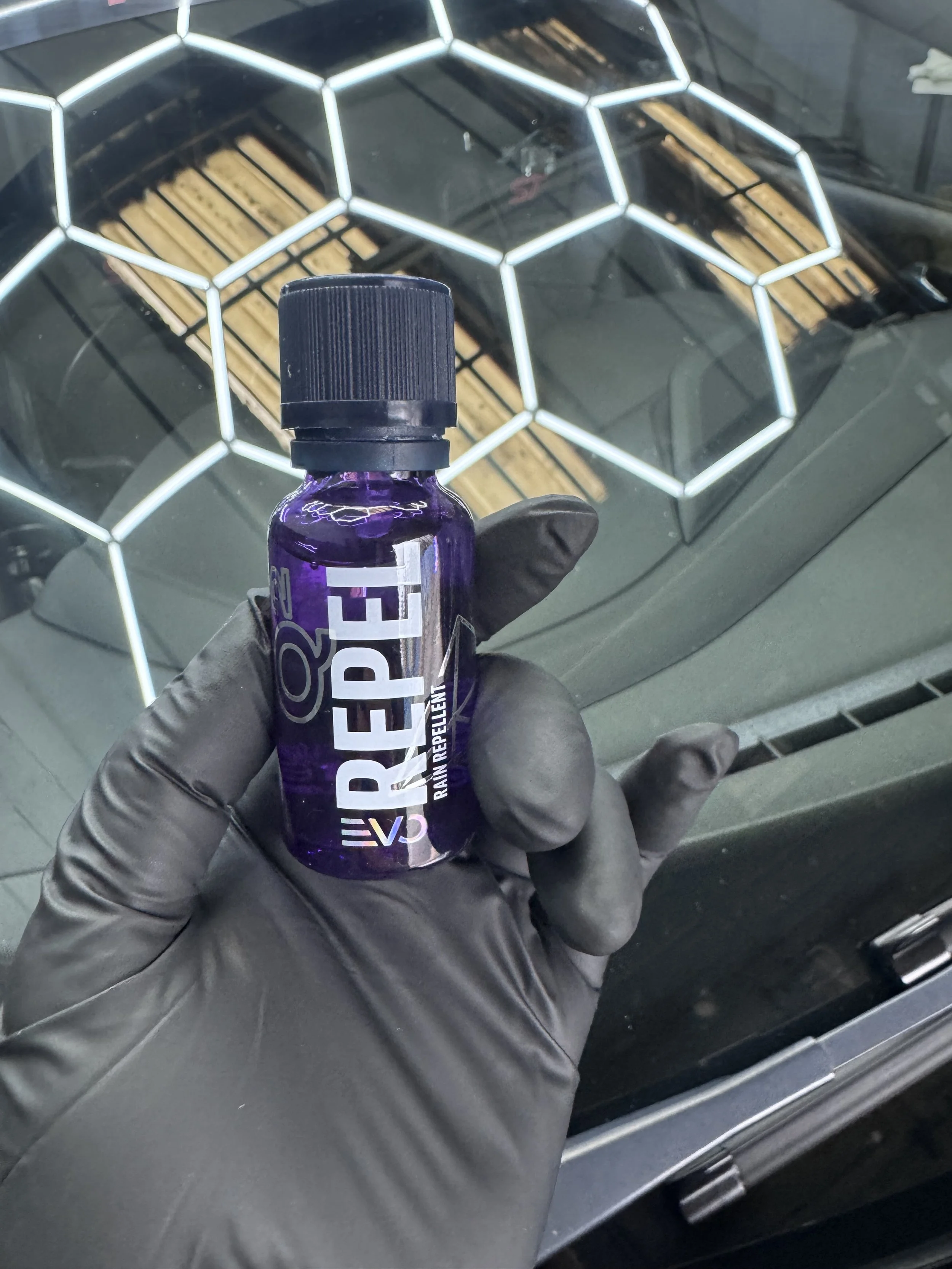 A person wearing a black glove holding a small purple bottle labeled 'Reepez' in front of a car's windshield with a honeycomb-patterned grille visible behind the glass.