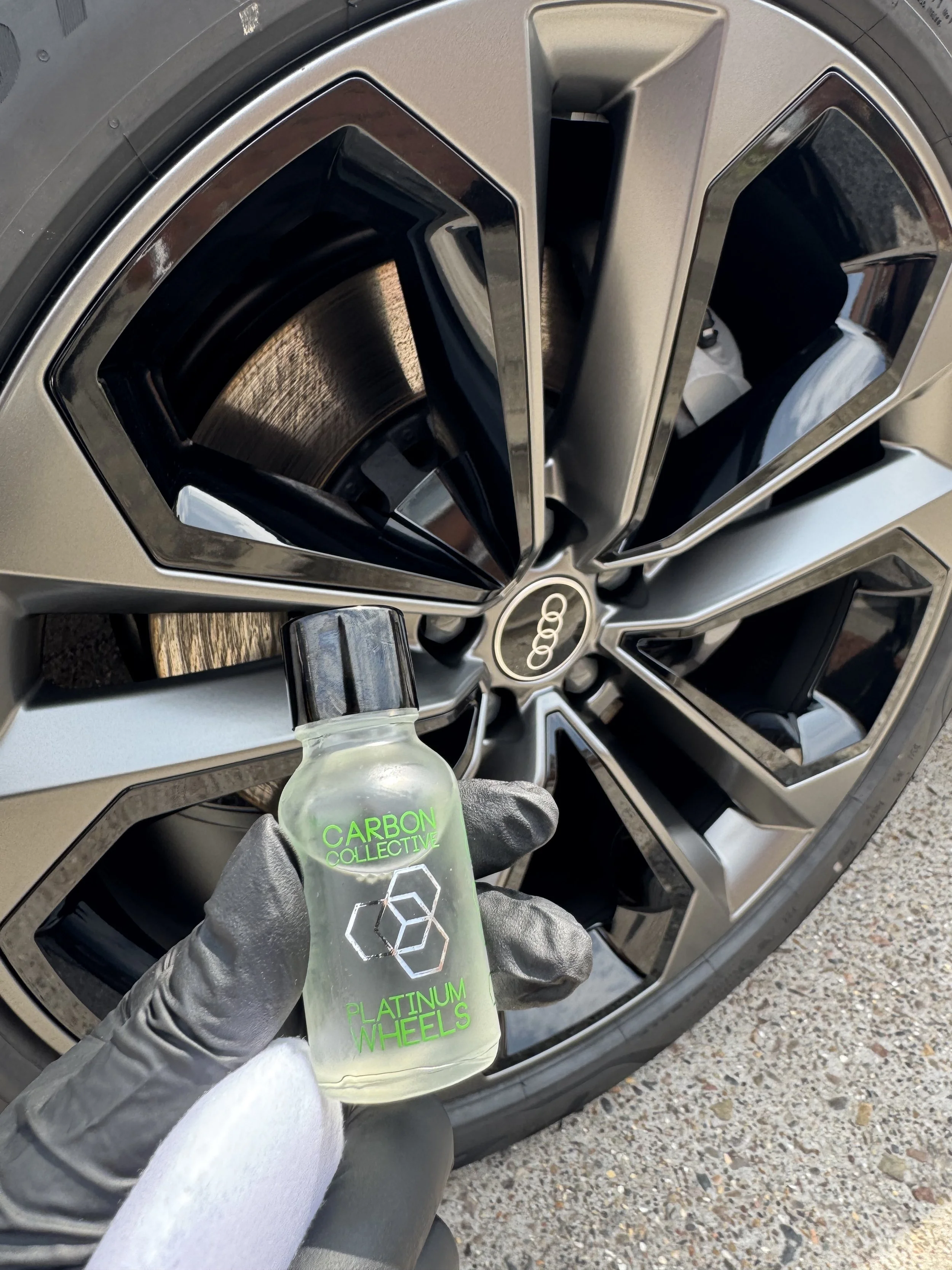 Close-up of a hand wearing a black glove holding a small bottle of Plasma Wheels tire cleaner positioned in front of an Audi wheel with a distinctive multi-spoke design.