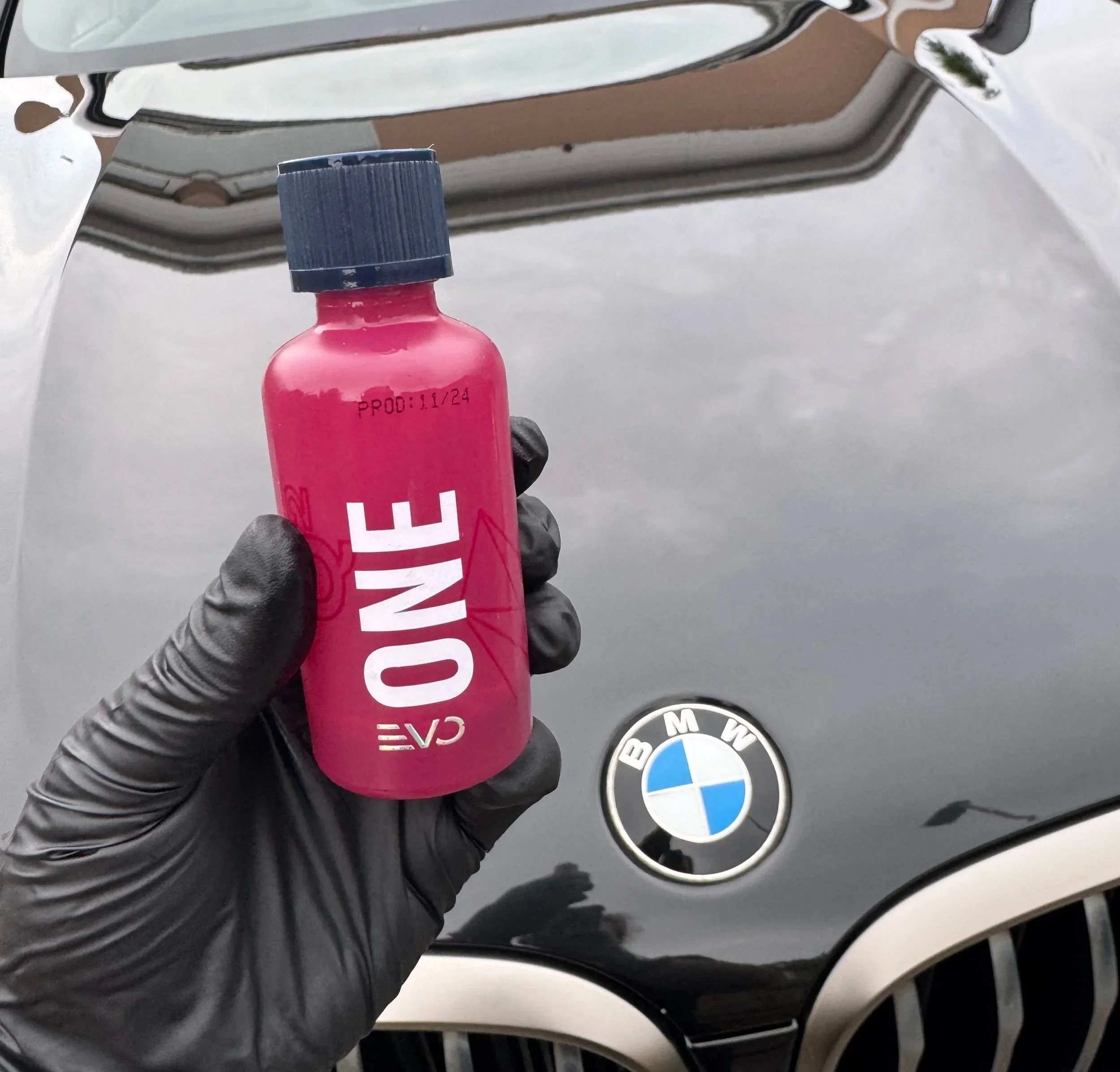 A person wearing a black glove is holding a small pink bottle labeled 'ONCE' in front of a black BMW car. The BMW logo is visible on the car's hood.