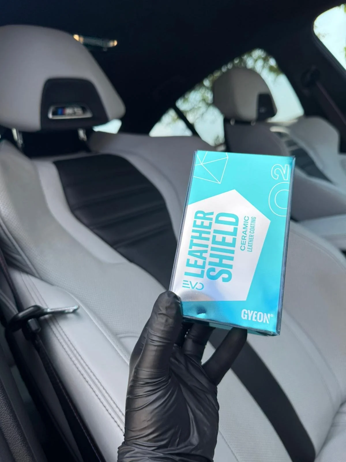 A hand wearing a black glove holding a box of Leather Shield ceramic leather coating in a car interior with light-colored leather seats and headrests.