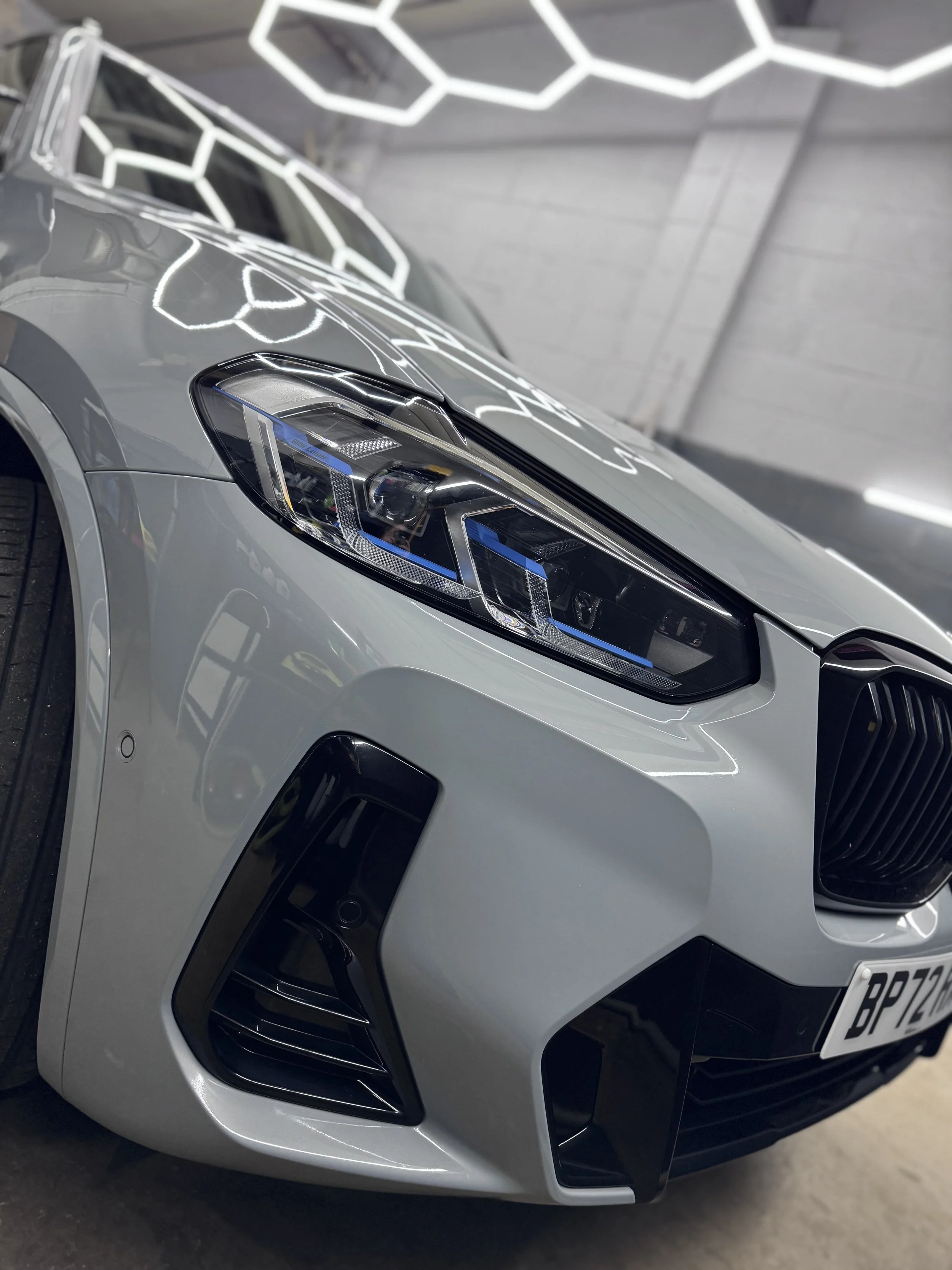 Close-up view of the front of a silver Audi car, showing the headlight, front grille, and part of the hood, with reflections of light fixtures on the car's surface.