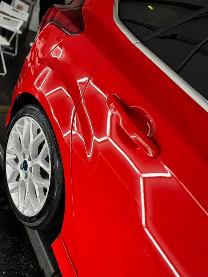 Close-up of the rear side of a shiny red car showing a white wheel with a black tire and the car's sleek body reflecting overhead lights.