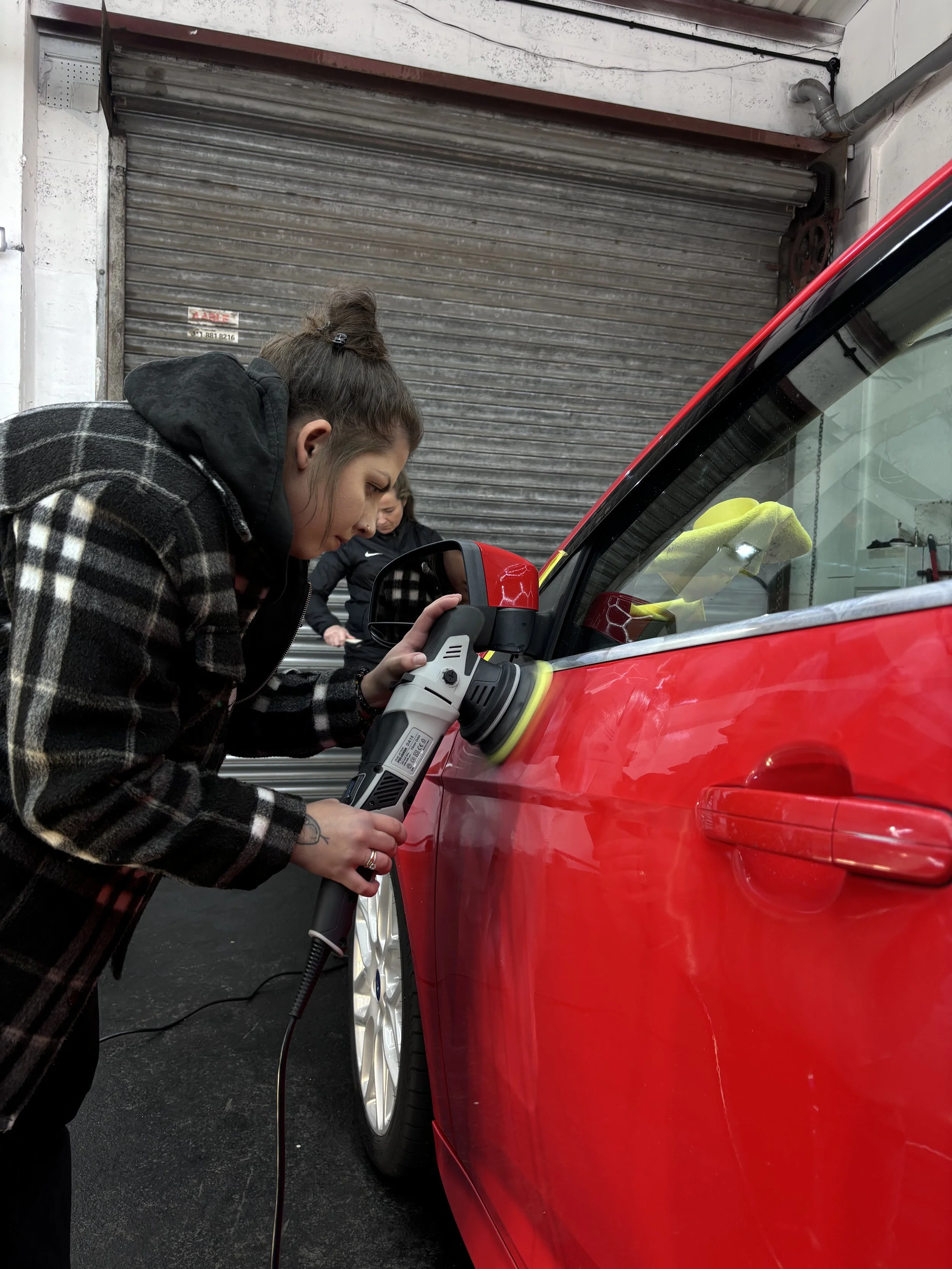 A woman in a black and brown plaid jacket uses a power buffer on the side of a red car in a garage with a metal roll-up door in the background.