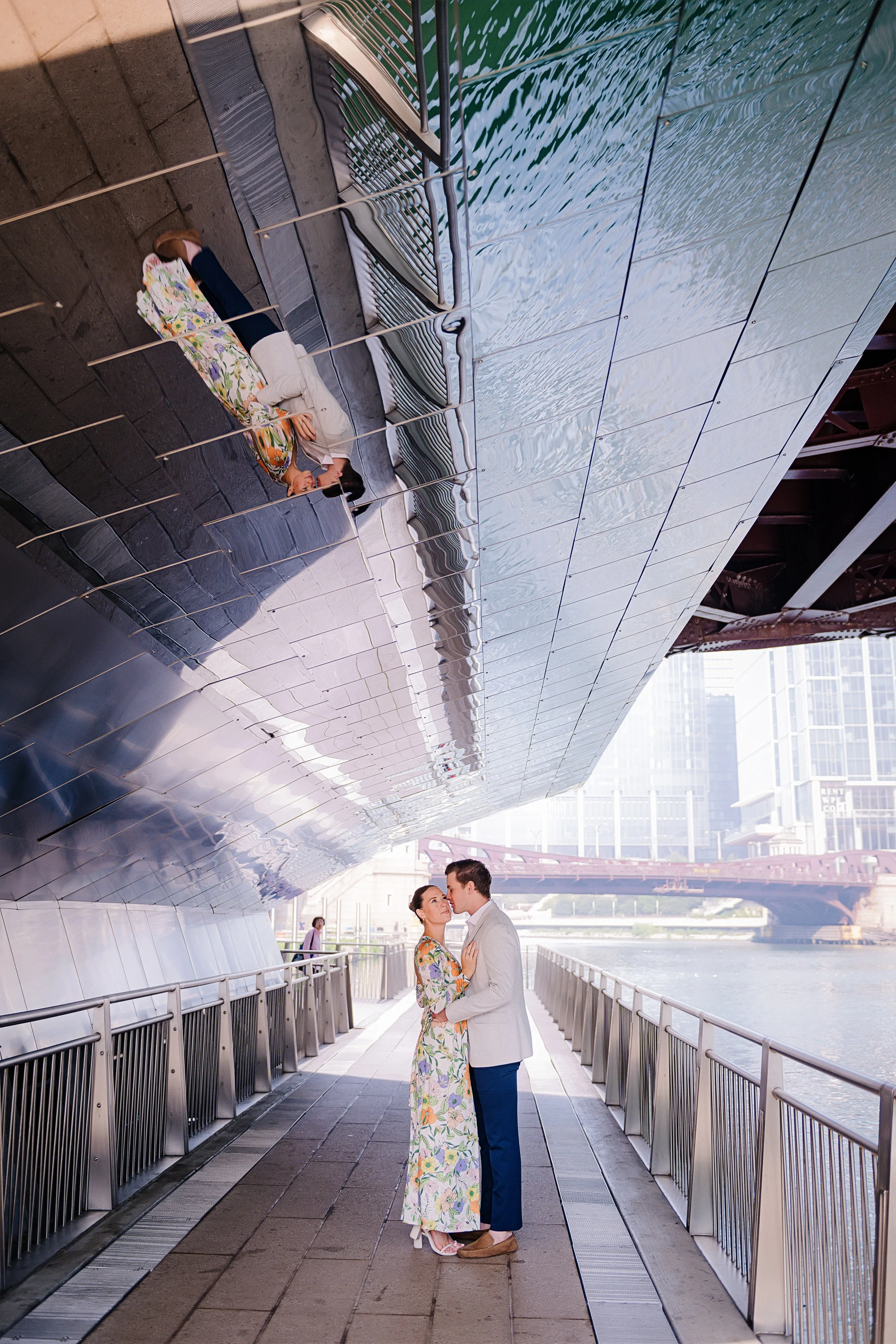 A couple stands close together kissing on a walkway by a river with a modern cityscape in the background. An upside-down reflection of the woman is seen on the metallic underside of a bridge.
