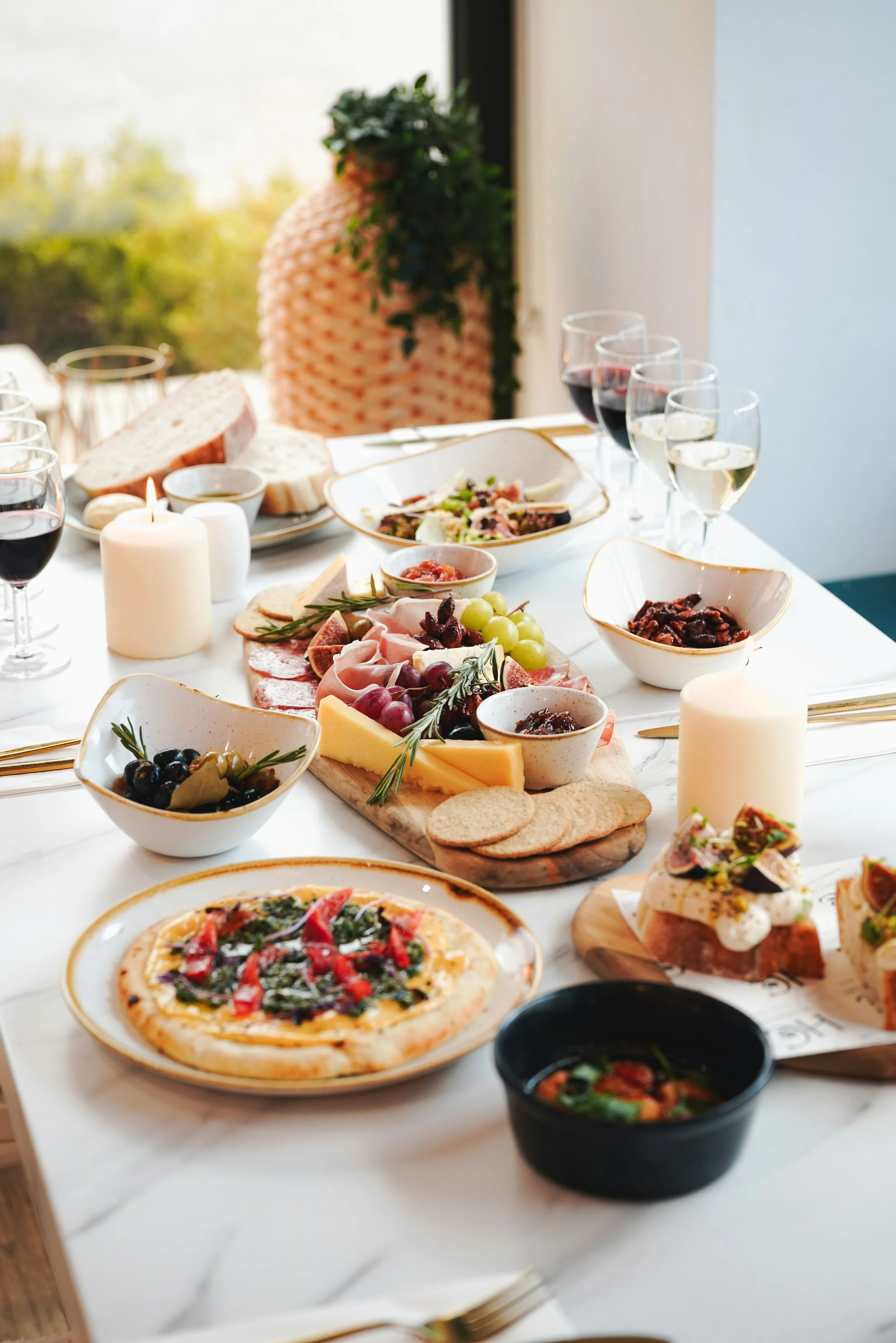A table set for a meal with various dishes including pizza, cheese, grapes, olives, bread, and charcuterie, along with wines and candles, in a bright room with a large window in the background.