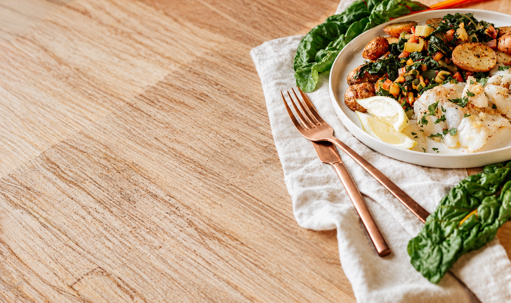 A white oval plate with cooked fish fillets garnished with herbs, lemon wedges, roasted potatoes, and a vegetable medley, on a wooden table with a white cloth and copper utensils.