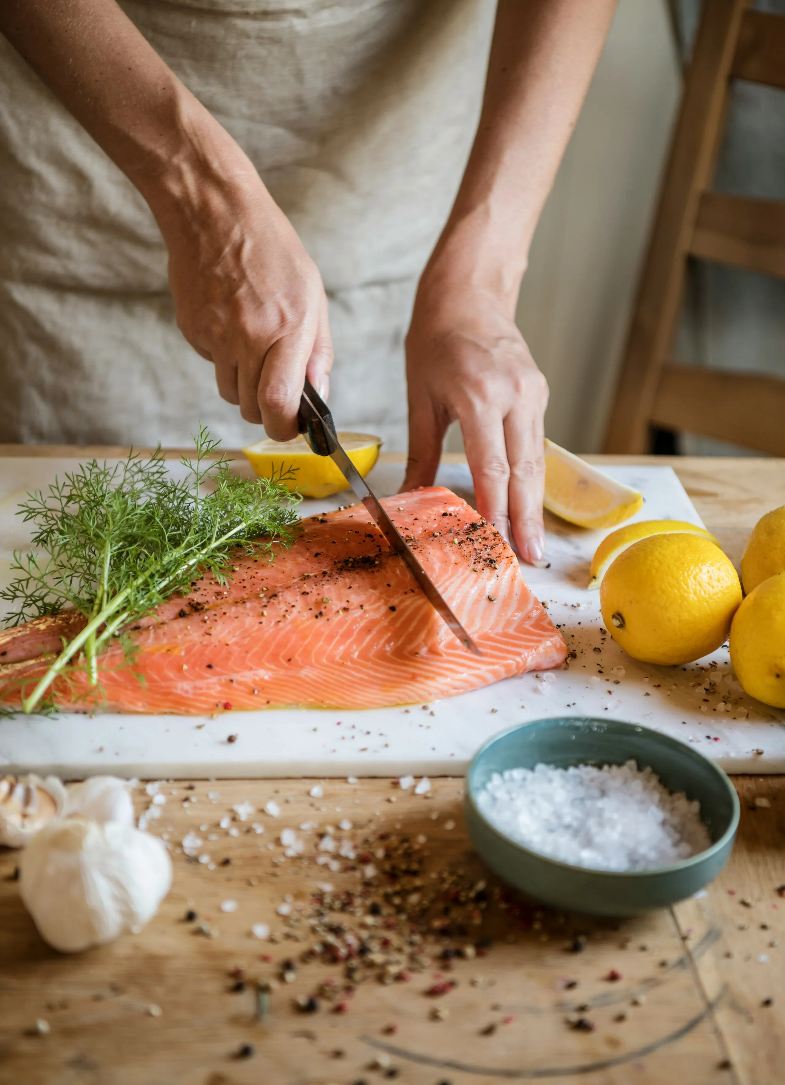 Person slicing raw salmon on a cutting board with fresh lemon slices, herbs, pepper, and salt nearby in a rustic kitchen.