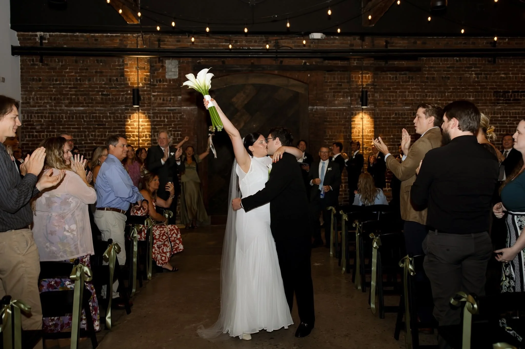 Wedding ceremony with a bride and groom kissing, surrounded by guests clapping and celebrating inside a rustic venue with brick walls and string lights.