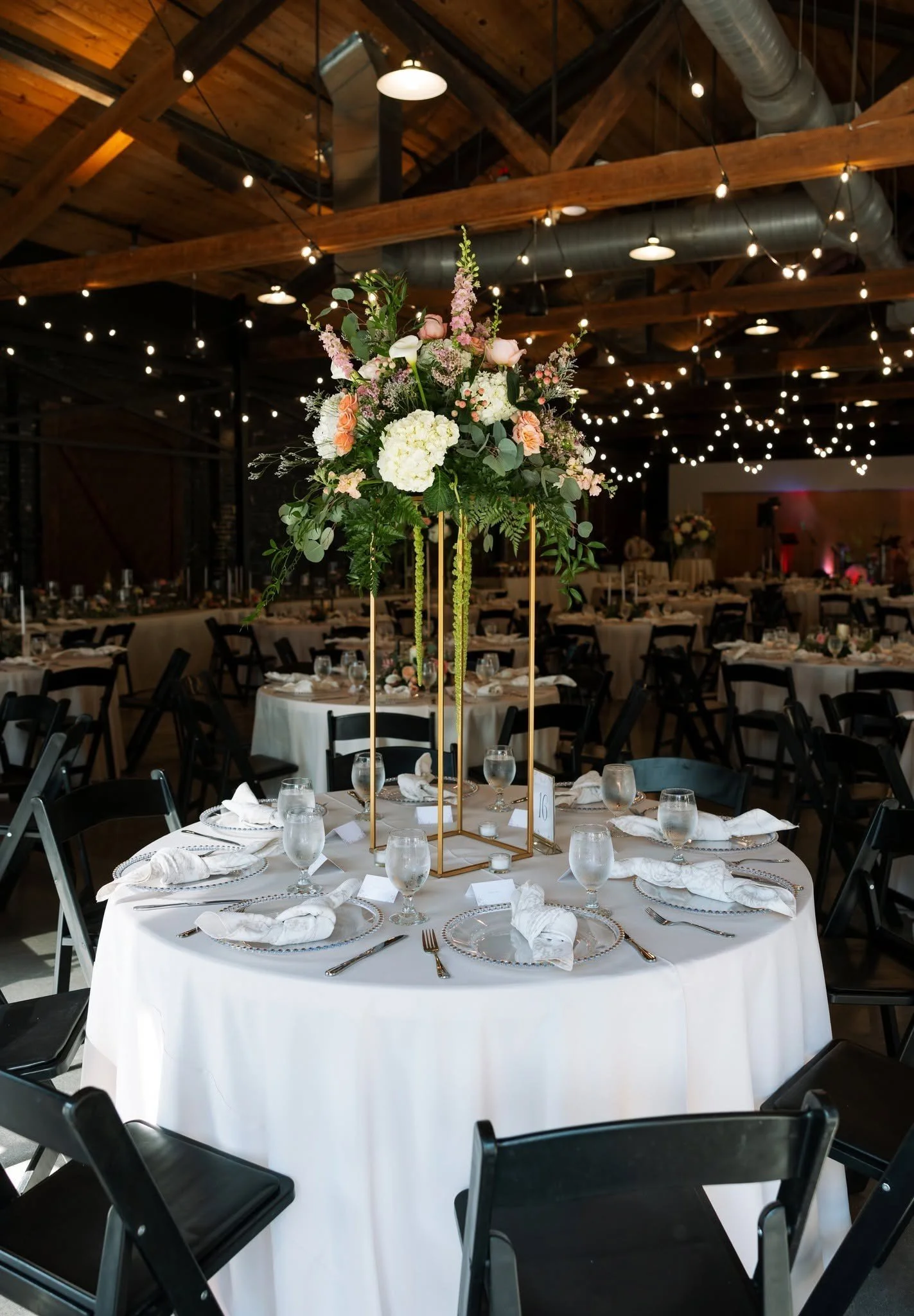 A round banquet table covered with a white tablecloth, set with silverware, glasses, and napkins. The centerpiece is a tall floral arrangement with pink and white flowers in a gold geometric stand. The background shows a rustic venue with wooden beams, string lights, and additional tables.