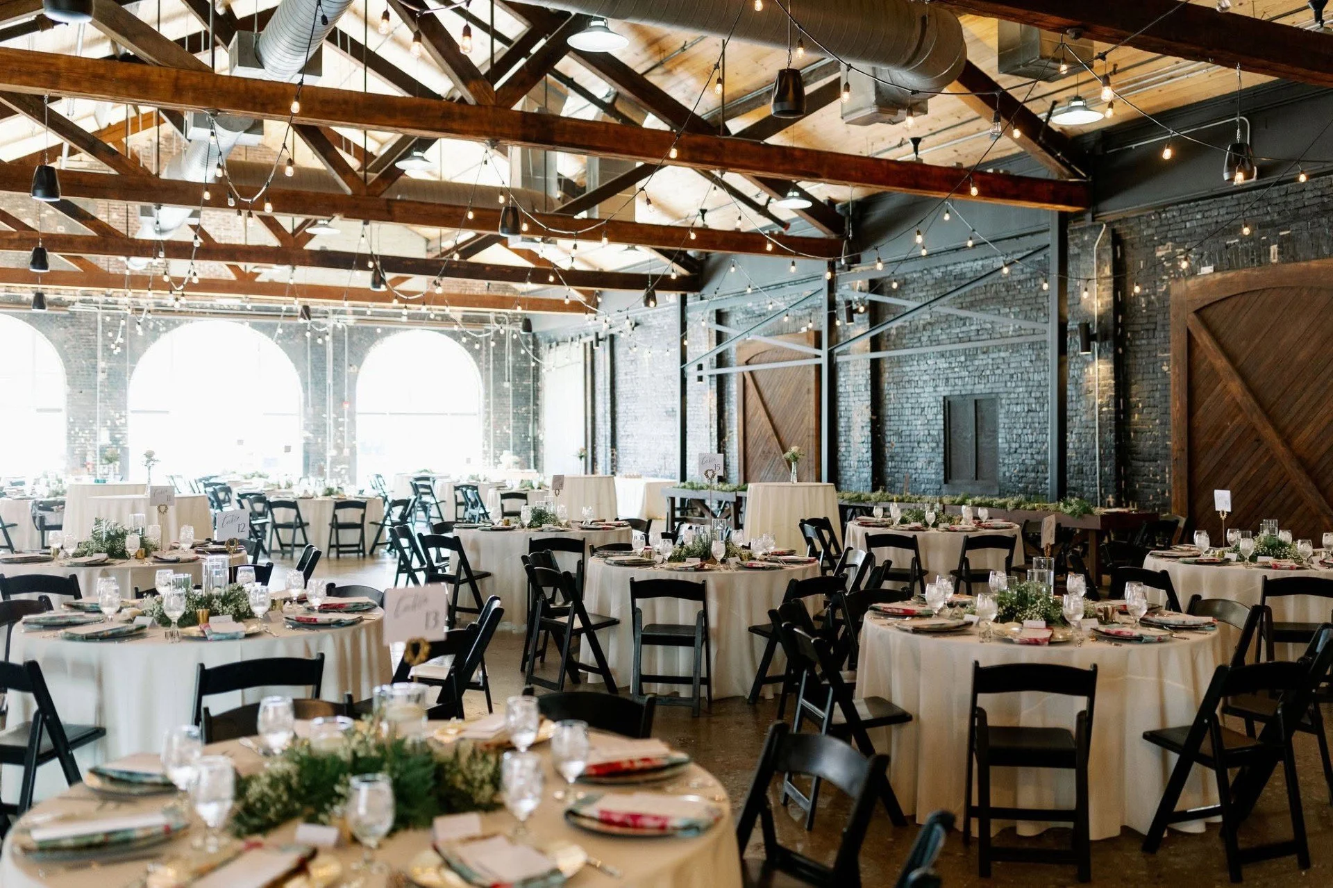 Interior of a rustic wedding reception hall with exposed brick walls, wooden beams, string lights, and decorated round tables with white tablecloths, glassware, and centerpieces.