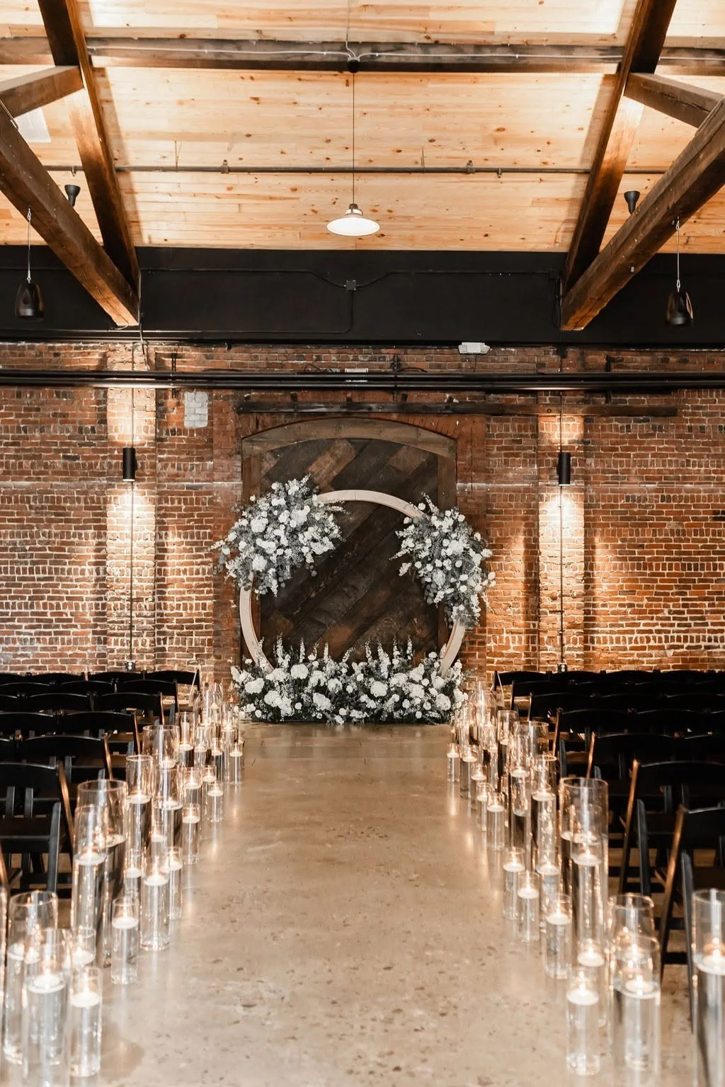 Indoor wedding ceremony setup with chairs, candles, and floral decorations against a brick wall and wooden ceiling.