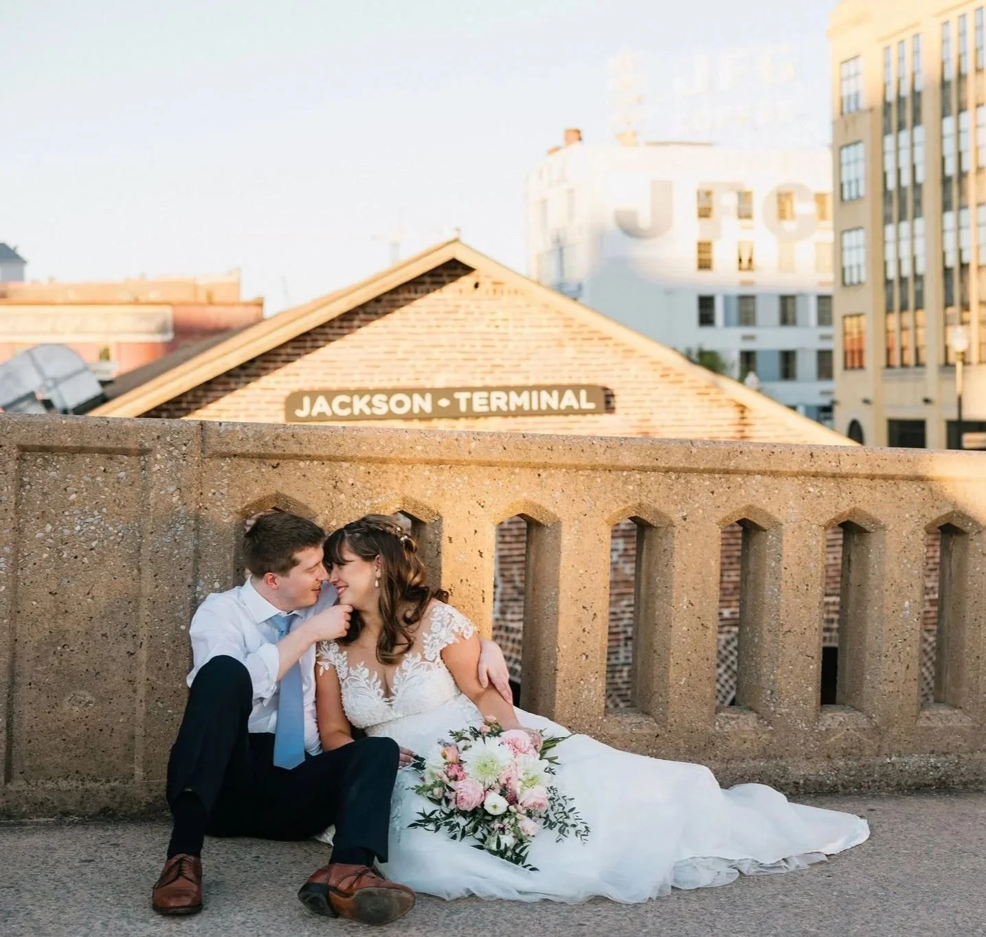 A newlywed couple sitting close together, smiling and touching foreheads, in front of a brick wall and a sign that reads "JACKSON TERMINAL" on a city street.