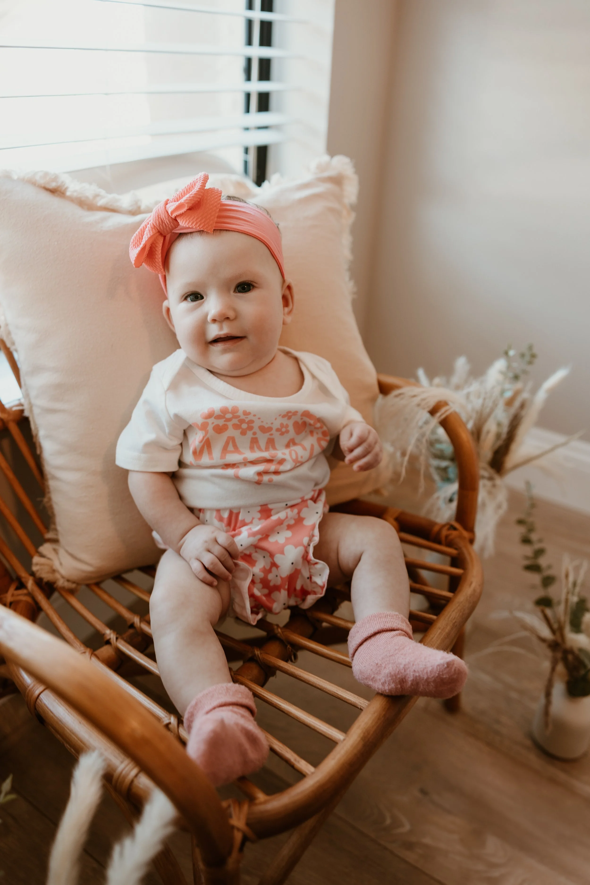 A baby girl sitting on a woven rattan chair near a window with blinds, wearing a pink headband with a bow, a white shirt with pink lettering, pink floral shorts, and pink socks.