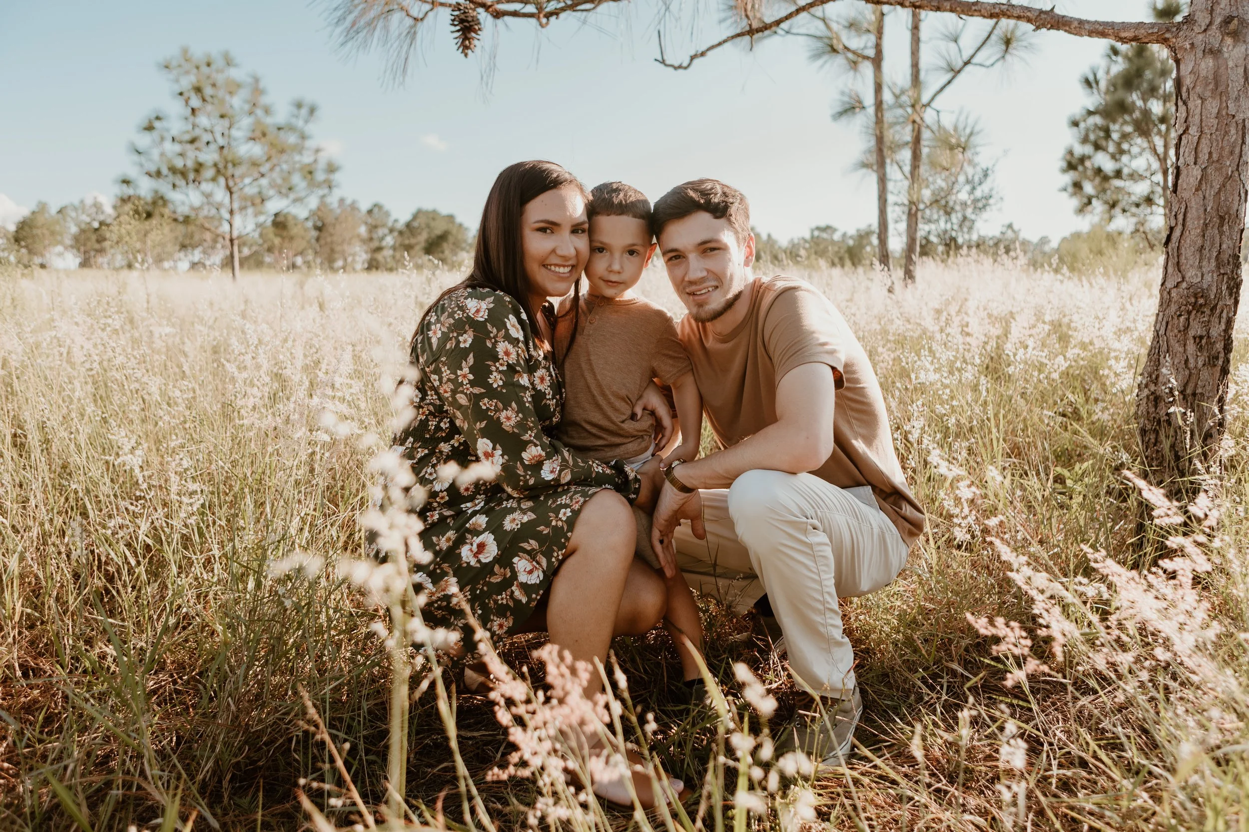 A family of three, a woman, a man, and a young boy, posing together in a grassy field with trees in the background on a sunny day.