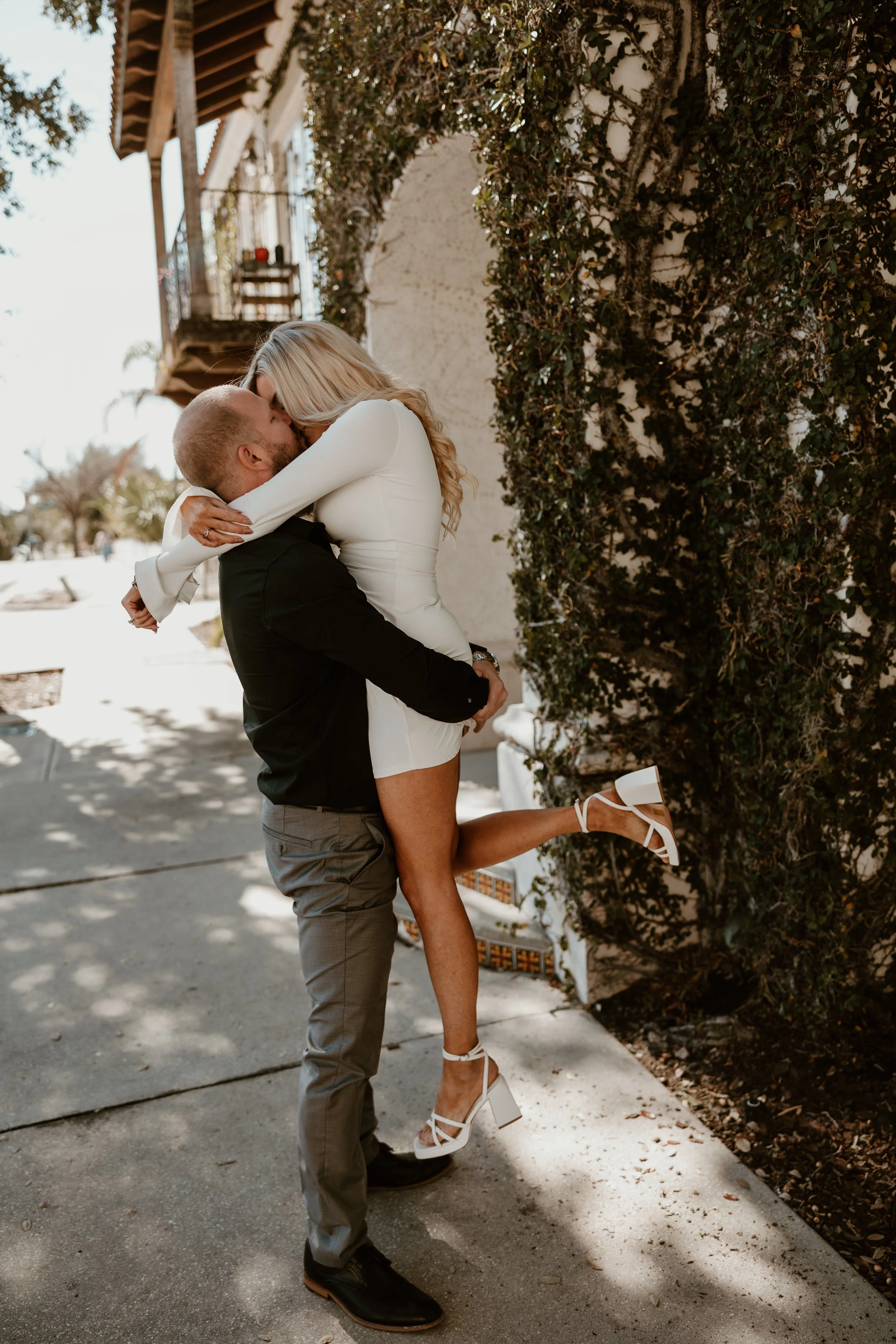 A man lifts a woman, both dressed formally, outside on a sunny day, sharing a kiss with the woman wrapping her arms around the man's neck and her legs around his waist.