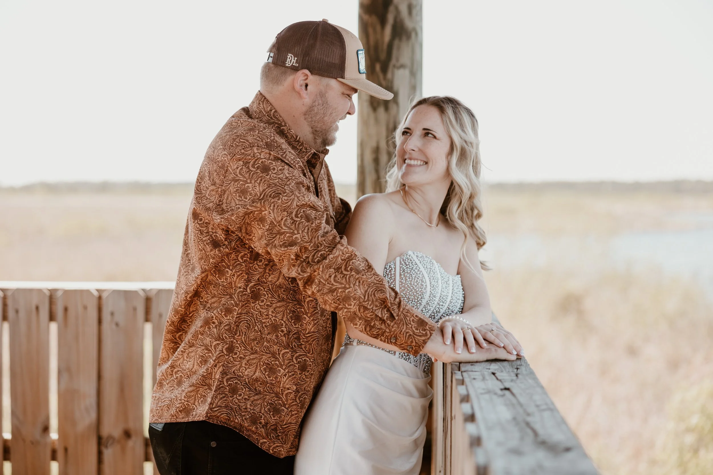 A happy couple leaning on a wooden railing outdoors, with the woman wearing a strapless beaded dress and the man in a patterned shirt and cap, smiling at each other.