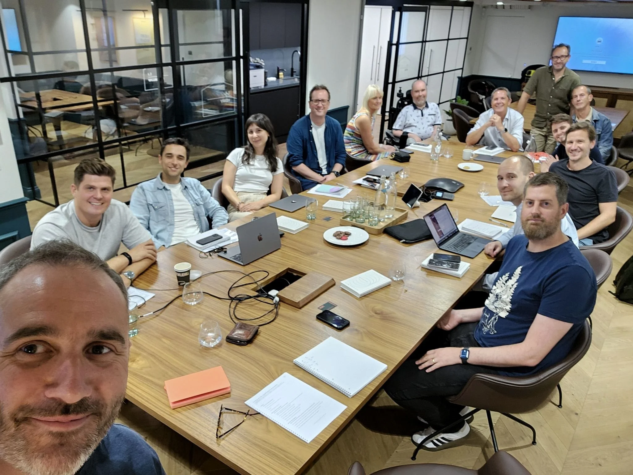 A group of people in a business meeting around a conference table, with laptops, notebooks, and drinks. One person takes a selfie. The room has glass walls and a large screen on the wall.