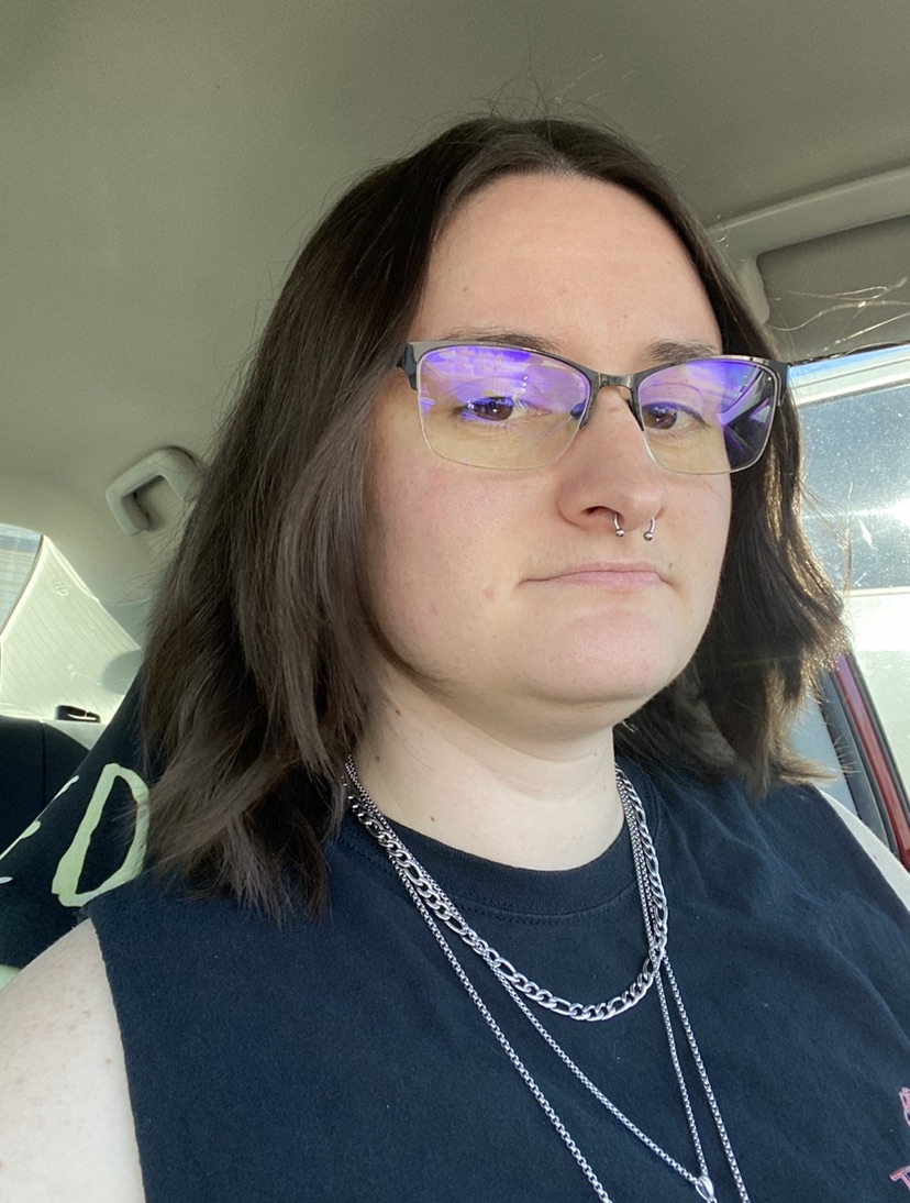 A person with shoulder-length brown hair and light skin wearing glasses, multiple silver necklaces, and a black sleeveless shirt sitting inside a vehicle.