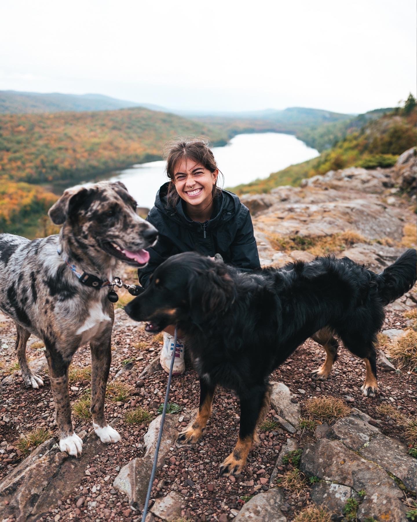 Young woman smiling with two dogs on a rocky trail with a scenic view of a lake and forested hills in the background.