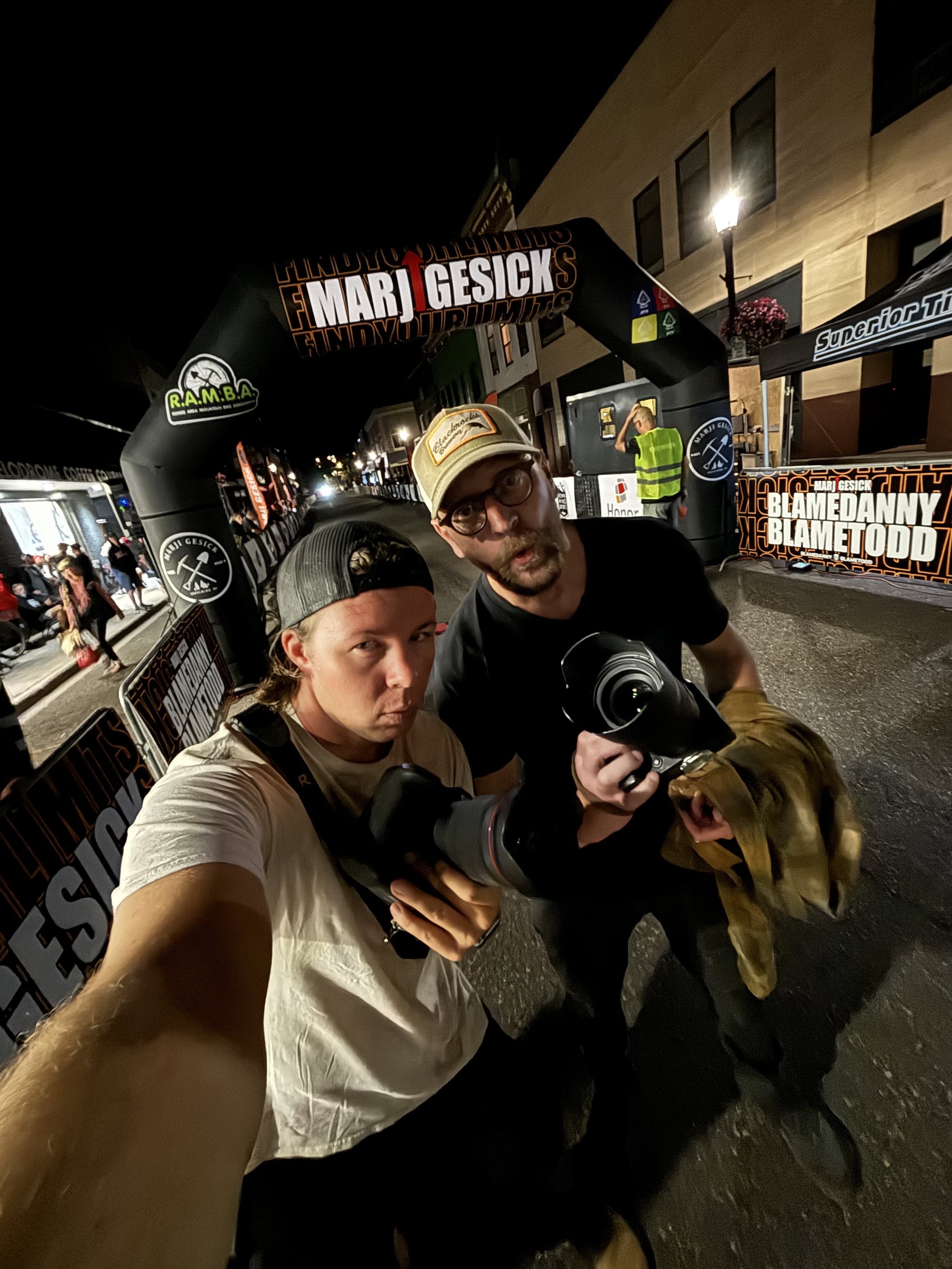 Two men taking a selfie at night in front of an event entrance with signs for Marji Gesick race. One holds a camera and wears casual clothing.