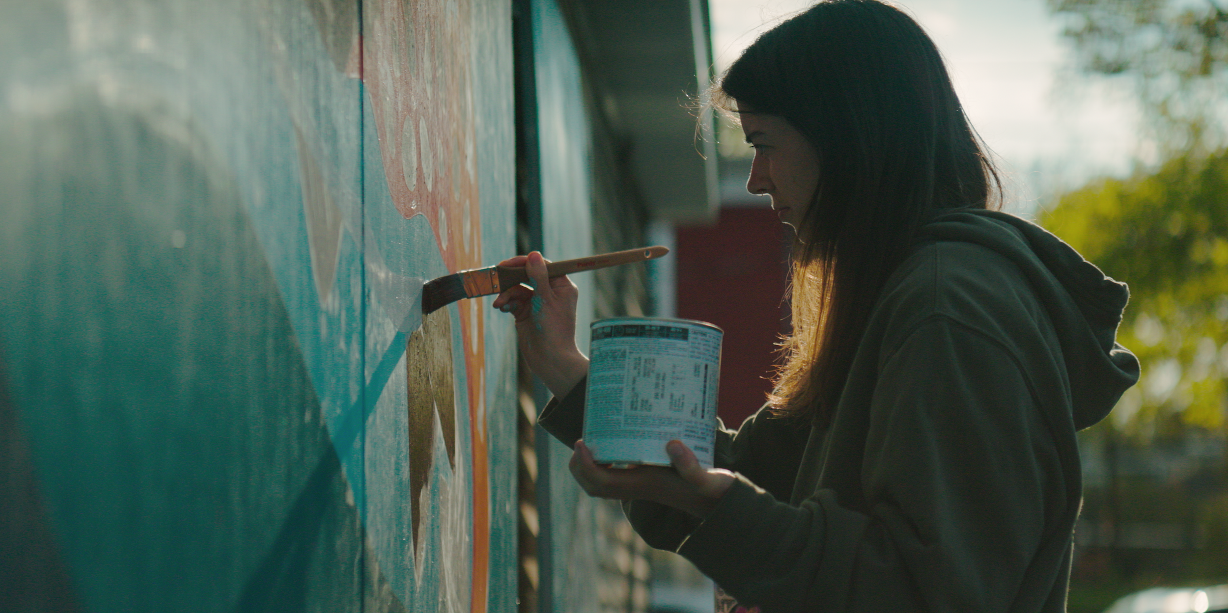 Person painting a mural with a paintbrush, wearing a hoodie, outdoors.