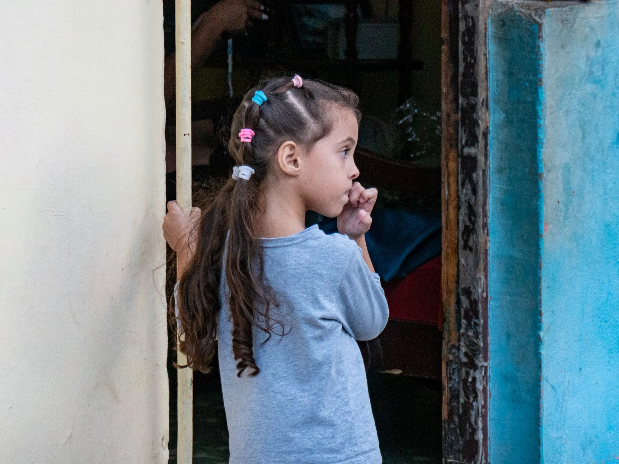 A young girl with long, curly brown hair styled with colorful hair ties, wearing a gray long-sleeve shirt, standing in profile, looking thoughtfully into an opening between a cream-colored wall and a wooden doorframe - Photograph by Noemi Moreno