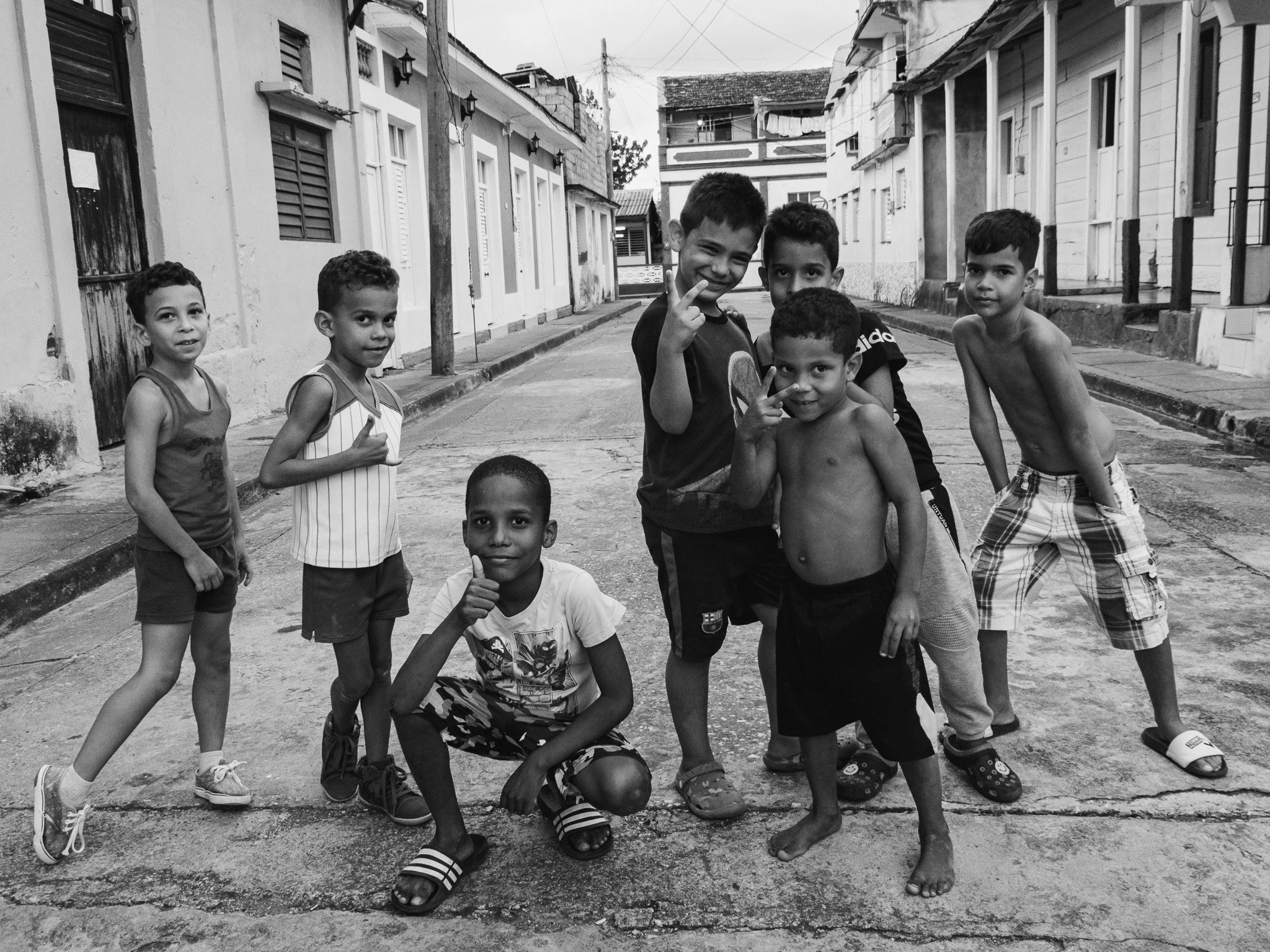 Group of seven boys standing and squatting on a street in a neighborhood, some making peace signs, with houses in the background.